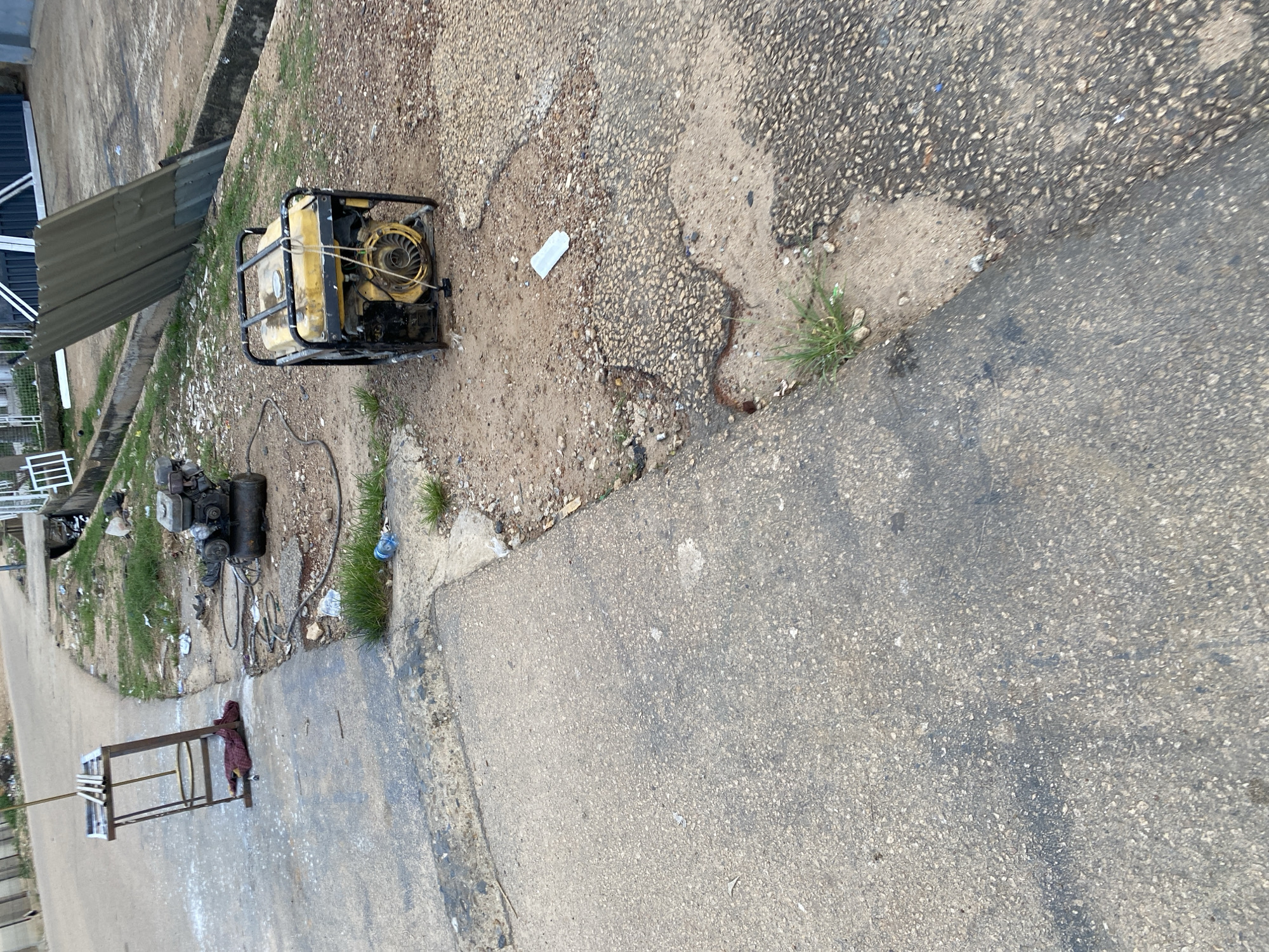 This image shows an outdoor scene in a city that appears to be Jos, Nigeria. The foreground is dominated by a rough, grey concrete surface that shows signs of wear and tear, with darker patches and scattered debris. To the right, the concrete transitions into a patch of dirt and gravel, also littered with small pieces of trash.
In the mid-ground, two generators are visible. One is a dark, older model with an engine and a tank, situated on the dirt/gravel area. It is connected to cables that snake across the ground. To its right, a yellow and black framed generator stands on the same patch of dirt. It has a coiled cord hanging from its front and a pull-start mechanism.
Further back, a metal stand with a red cloth draped over it is positioned on the concrete. It appears to be a makeshift structure, possibly for holding materials. Beyond this, a paved road curves away, bordered by a concrete drainage ditch. The ditch contains debris, and a large, corrugated metal sheet is propped up against a fence in the background. The overall environment suggests a working area or a place where equipment is stored, possibly in a less maintained part of the city. The presence of the generators and the stand implies some form of mechanical work or an electrical service is being provided or has recently occurred.