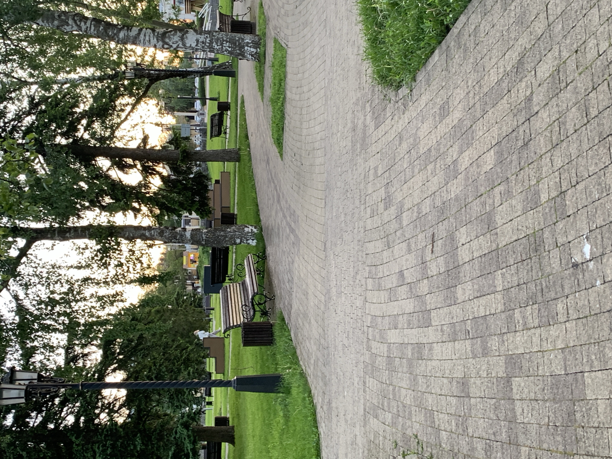 The image shows a walkway in a park in Stavropol, Russia. The walkway is paved with gray bricks and curves through the park. Benches line the walkway, and trees provide shade. A lamppost is visible near the bottom left. In the distance, there are other trees and possibly headstones, suggesting this area might be near a cemetery or memorial garden. The sky appears to be illuminated by the setting sun. A person is sitting on one of the benches.