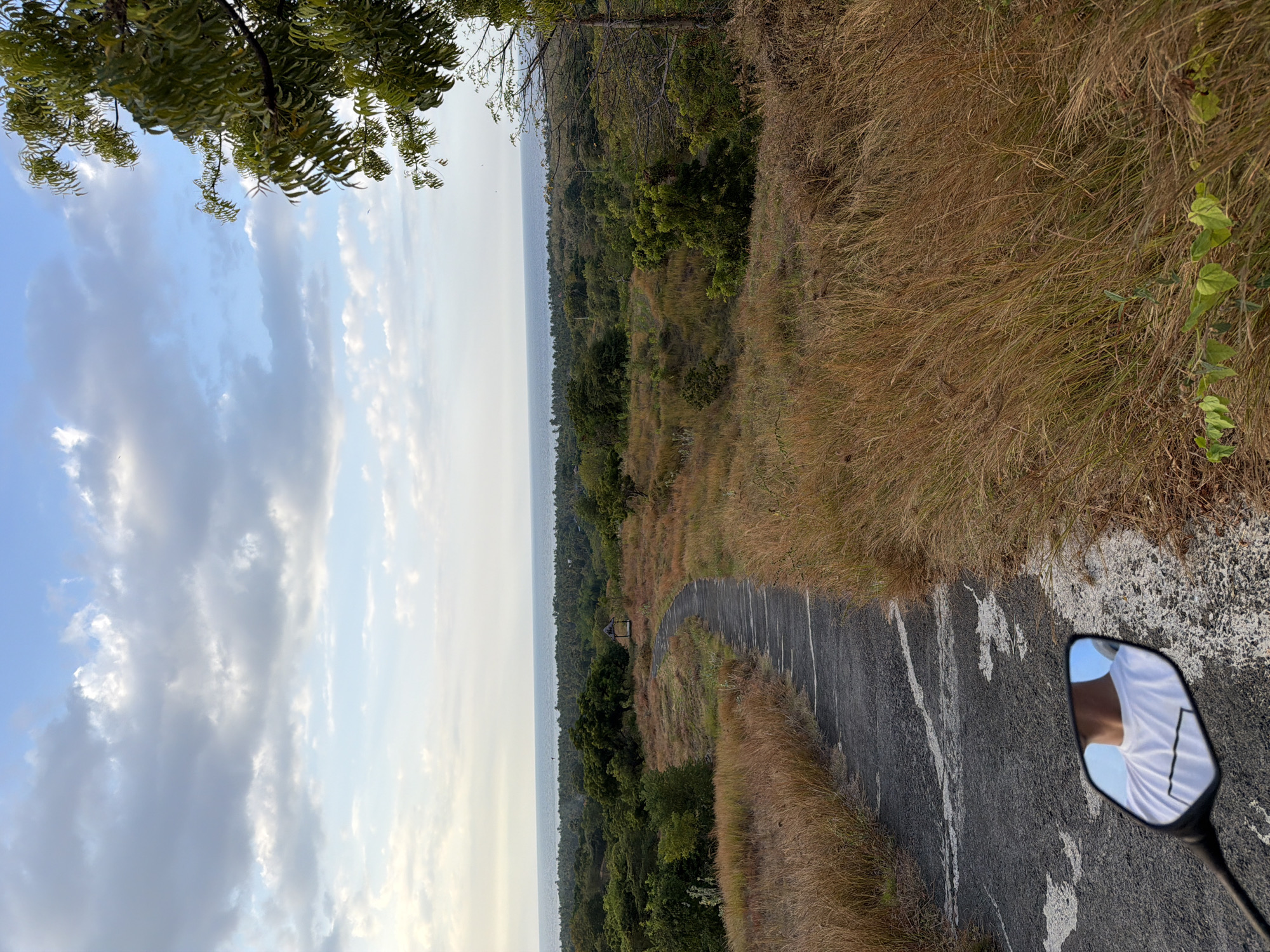 The image captures a scenic view of a paved road winding through dry, golden grass and sparse trees, leading towards the ocean horizon. A small, open-sided shelter is visible in the middle distance, nestled among the greenery. The sky is a mix of blue with scattered, light gray clouds. In the foreground, a motorcycle's side mirror reflects a person wearing a white t-shirt with a dark rectangular graphic. The scene suggests an outdoor location, possibly a viewpoint or a rural area on the outskirts of Kubutambahan, Indonesia, given the vegetation and coastal proximity. The lighting indicates it is either early morning or late afternoon.