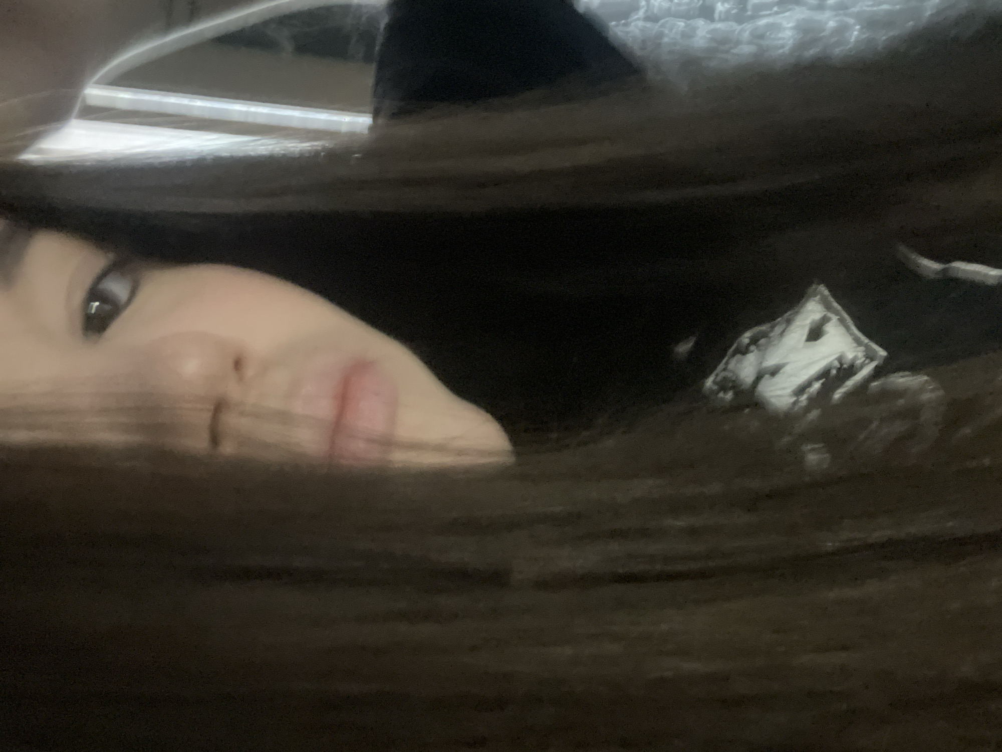 The image displays a close-up portrait of an individual in an indoor setting within the city of Chaikovsky, Russia. The subject's face is partially obscured by long, dark brown hair that falls across the left side of the frame, covering the forehead and upper left portion of the face. The visible features include the individual's right eye, nose, and mouth. The right eye is dark and appears to be looking towards the viewer. The nose and mouth are central, with lightly colored lips. The individual is wearing a dark-colored garment, possibly a t-shirt or sweatshirt, with a partially visible white graphic print on the lower left side of the chest area. This graphic is indistinct but appears to be an image or symbol. In the background, on the right side, a light-colored, possibly textured or knitted object is discernible, suggestive of a blanket, pillow, or furniture upholstery. Reflections or light sources are subtly visible in the upper right background. The scene depicts a static pose with no discernible actions or interactions beyond the subject looking towards the camera.