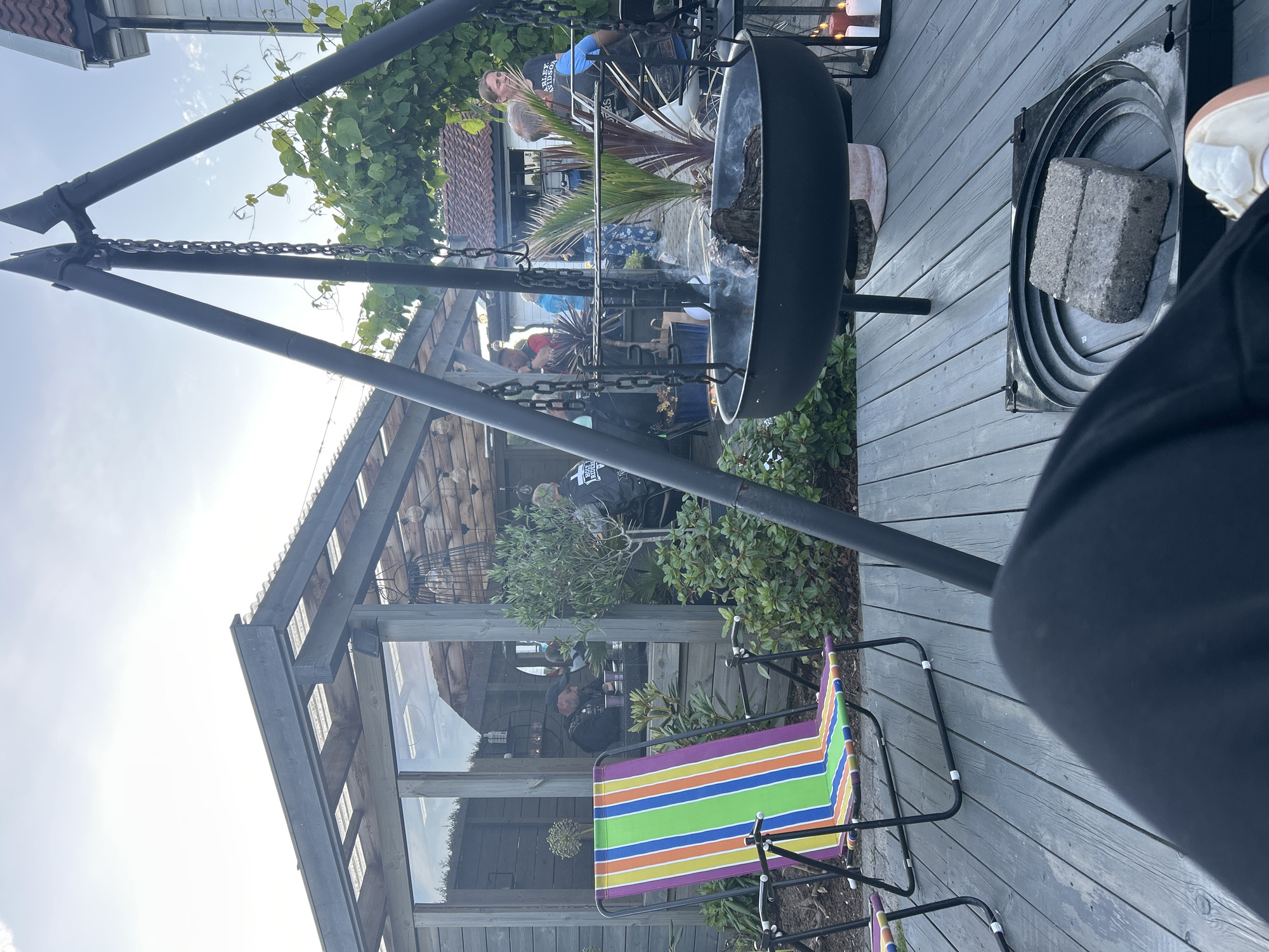 The image depicts an outdoor gathering scene at a private residence in Sandefjord, Norway, likely during a summer evening. A group of people, some wearing motorcycle club attire, are socializing around a fire pit. The environment includes a wooden deck, a greenhouse structure with glass walls, lush green vegetation, and outdoor furniture.
Specifically, a large black fire pit hangs from a tripod structure, with smoke emanating from burning wood within. In the foreground, a person's legs are visible, with a colorful striped deck chair to their left. Behind the fire pit, several individuals are engaged in conversation. One woman, wearing a black t-shirt with "HARLEY-DAVIDSON" printed on it, is looking to the right. Another man in the background is wearing a black vest over a t-shirt.
The greenhouse, to the left of the frame, has wooden framing and transparent roofing, with people visible inside. The overall atmosphere suggests a relaxed and informal social event. The lighting indicates it is either late afternoon or early evening, with the sky overcast.