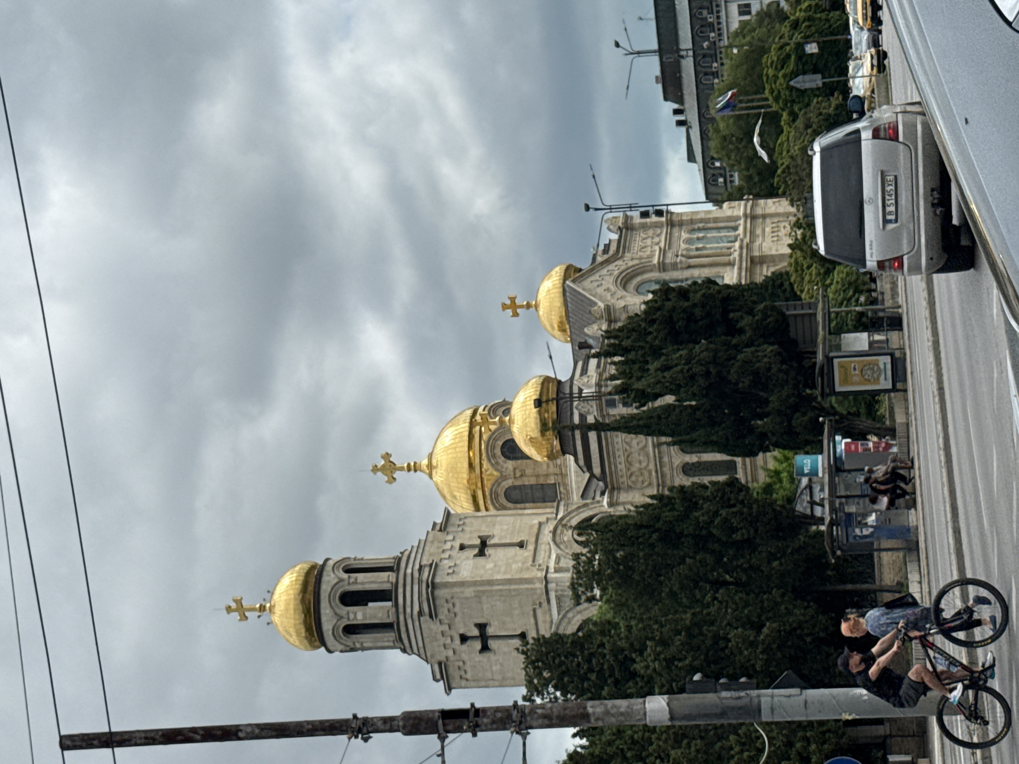 An outdoor, eye-level, medium shot captures the Assumption Cathedral in Varna, Bulgaria, under a cloudy sky. The grand cathedral, characterized by its prominent gold domes and cross-topped spires, dominates the upper half of the frame. In the foreground, a man wearing a black t-shirt and shorts cycles on a bicycle from left to right, his back to the camera. He is accompanied by a woman in a blue patterned dress walking alongside him. Across the street, a silver Mercedes SUV is visible, and beyond that, a bus stop with several people waiting. Trees line the street, and electrical wires are strung across the top of the image.