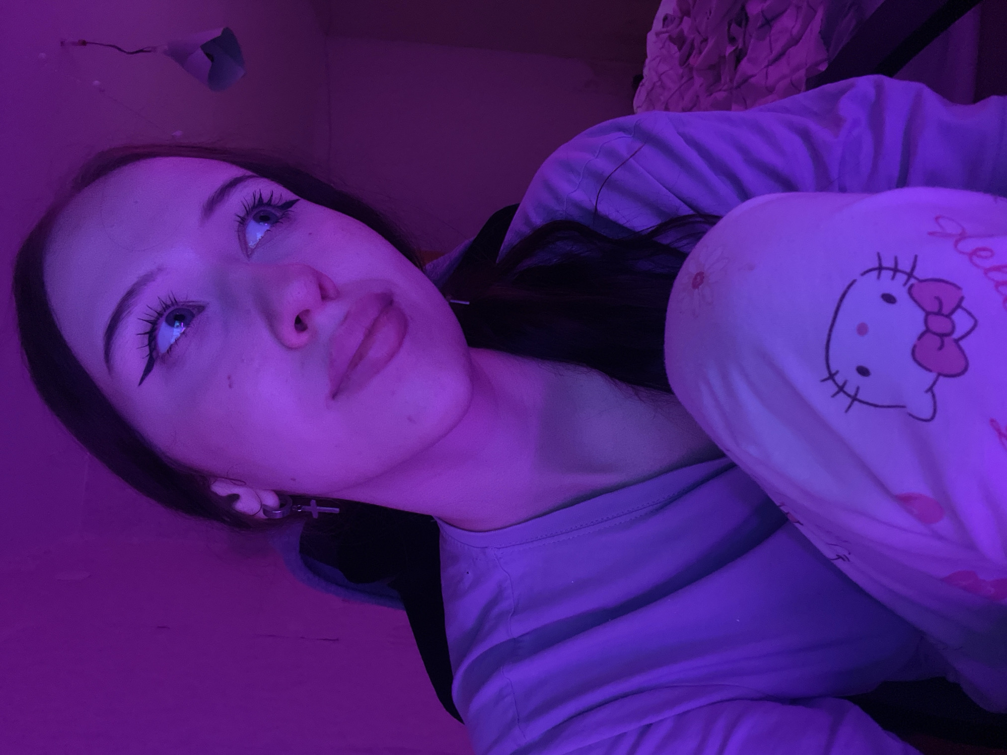 The media file is a close-up, portrait-oriented photograph of a young woman with long, dark hair. She is looking upwards and to her right, with her eyes wide open. Her makeup includes noticeable eyeliner and mascara. She is wearing a light-colored t-shirt and light-colored pants with a Hello Kitty pattern. The lighting in the image is predominantly purple, suggesting an artificial light source, possibly a colored bulb or LED. A portion of a patterned fabric, possibly bedding or a blanket, is visible in the upper right background. The woman is wearing earrings, one of which is a silver hoop with a cross charm. The overall impression is that of a casual, indoor setting, possibly a bedroom, illuminated by purple light. The image appears to be a selfie or a candid shot, capturing a moment of reflection or contemplation. No specific time of day or weather conditions can be determined, but the indoor setting and lighting suggest it is likely nighttime or a dimly lit room. There is no visible text other than the "Hello" written on the Hello Kitty pattern. The subject's expression could be interpreted as curious or introspective.
