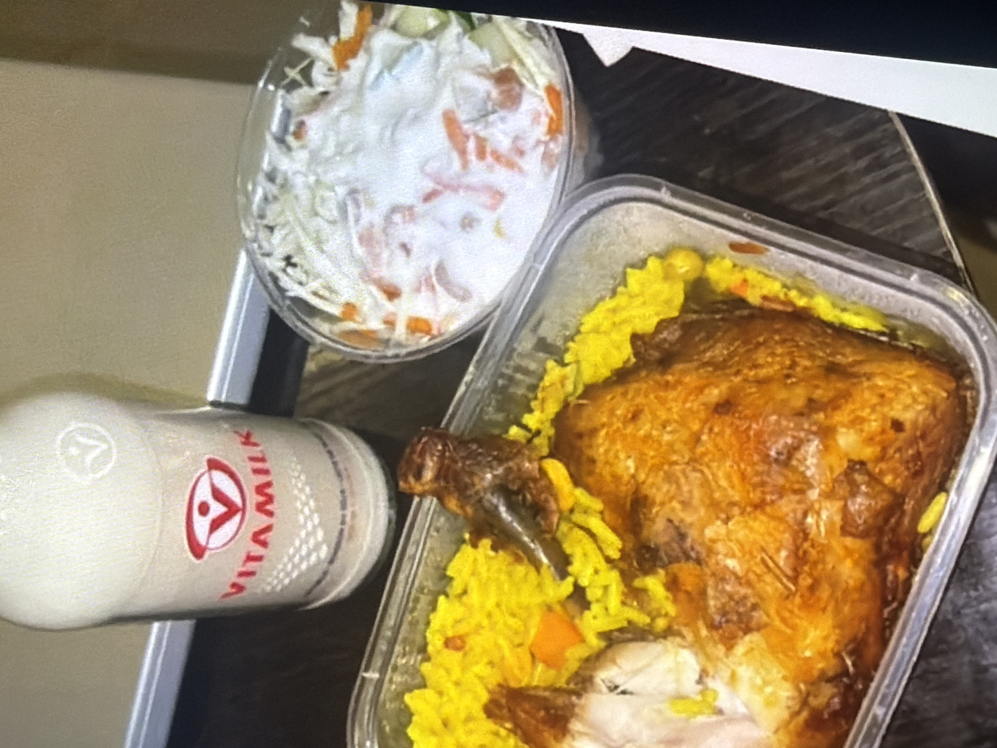 The image shows a meal with a bottle of Vitamilk, a container of coleslaw, and a container of roasted chicken with yellow rice. The Vitamilk bottle has a red and white logo. The coleslaw appears to be made with shredded cabbage, carrots, and a creamy dressing. The chicken is golden brown and looks crispy. The yellow rice is seasoned and has some pieces of carrot mixed in. The food is presented in plastic containers, suggesting it might be takeout or a casual meal. The background is a blurred beige wall. The image does not provide any information about the location in Bukuru, Nigeria.