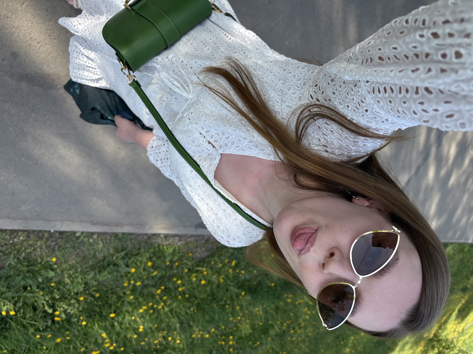 A woman wearing sunglasses and a white dress stands on a sidewalk with a grassy area to her left. She has a green bag slung over her shoulder and is sticking her tongue out. The grassy area contains yellow flowers. The location is not specified in the description, therefore it is not possible to identify the location as Izhevsk, Russia.