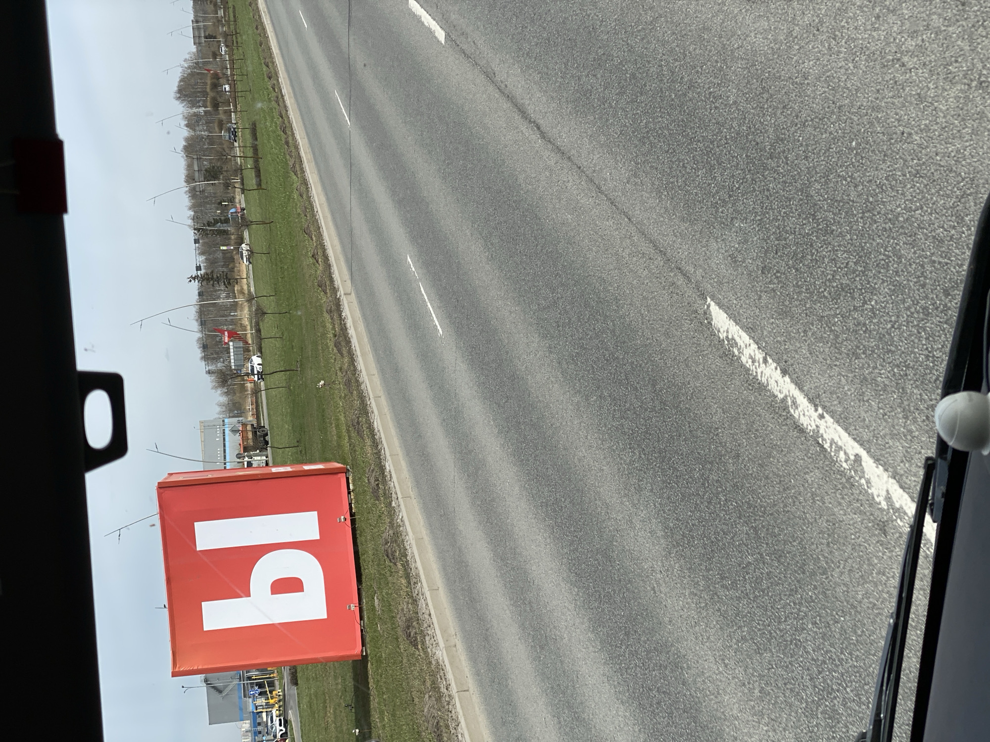 The image shows a view from a vehicle traveling on a road adjacent to a grassy area in Saint Petersburg, Russia. A large red advertising cube with white Cyrillic characters is visible on the side of the road. The road is asphalt and has white lane markings. There is a curb separating the road from the grass. Trees are visible in the background. The sky is overcast. The focus is on the roadside and the advertising cube.
