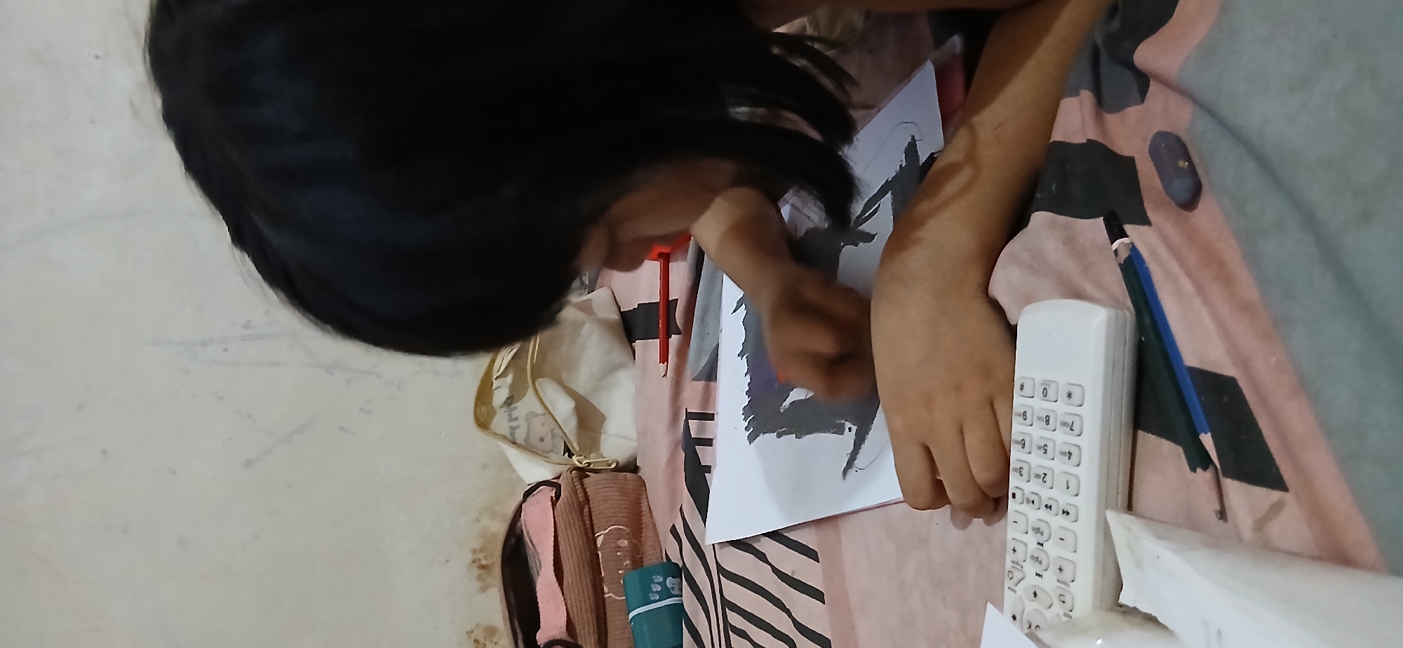 The image depicts a person with short black hair, seen from the side and slightly from behind, focused on drawing on a piece of white paper. The person's hands are visible, one holding a drawing tool, possibly a crayon or pastel, and the other supporting the paper. The drawing on the paper appears to be in dark shades, possibly charcoal or dark colored pencil, and depicts a somewhat abstract or silhouetted figure.
The scene is set indoors, likely in a bedroom or a similar personal space. The drawing is laid out on a surface covered with a patterned fabric, featuring diagonal stripes of pink, beige, and gray. In the background, there are two pouches or bags, one brown corduroy with a cartoon bear design and another cream-colored pouch with a teddy bear motif. A green cylindrical object with white markings is also visible to the left of the brown pouch.
In the foreground, a white remote control with numerous buttons is placed on the patterned fabric. To the right of the remote, a dual-colored pencil (blue and black) lies next to a small, dark purple eraser. A red pencil is also visible on the drawing paper, near the top edge. The wall behind the subjects is a pale, neutral color with some faint marks.
Given the context, this scene could be situated in any home in Samarinda, Indonesia. The objects and overall setting do not provide specific clues to pinpoint a precise location within the city.