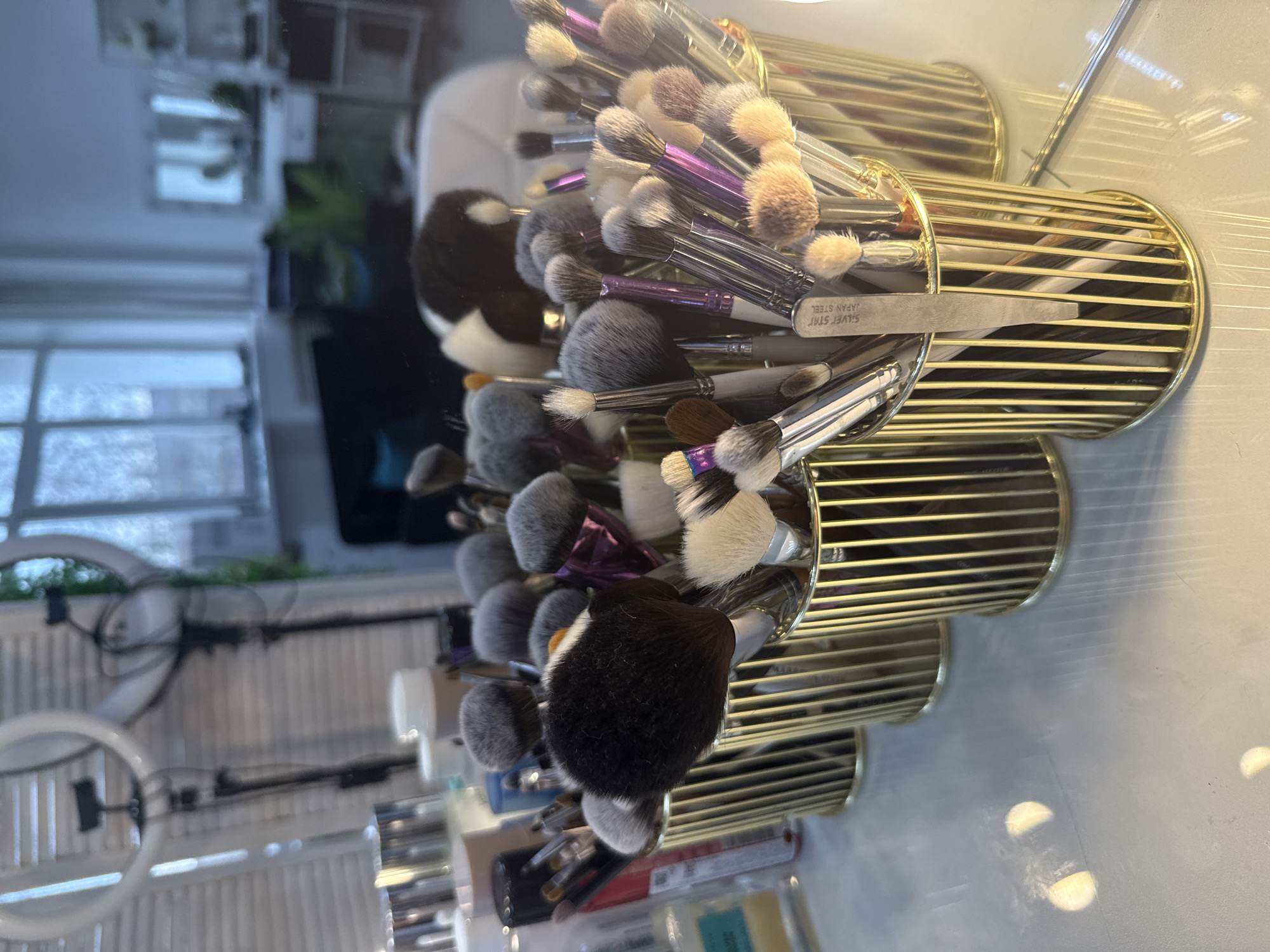 The image shows a collection of makeup brushes organized in gold-colored, cylindrical holders. The brushes have various bristle types and colors, with some having purple or silver handles. The holders are arranged on a white surface that appears to be reflective.
In the background, visible through a window, is an ambiguous urban scene. The presence of a ring light and other cosmetics items suggests this is a beauty setup, likely in a residential or studio setting. There are no visible people or clear identifiers to conclusively place the location in Chaikovsky, Russia.