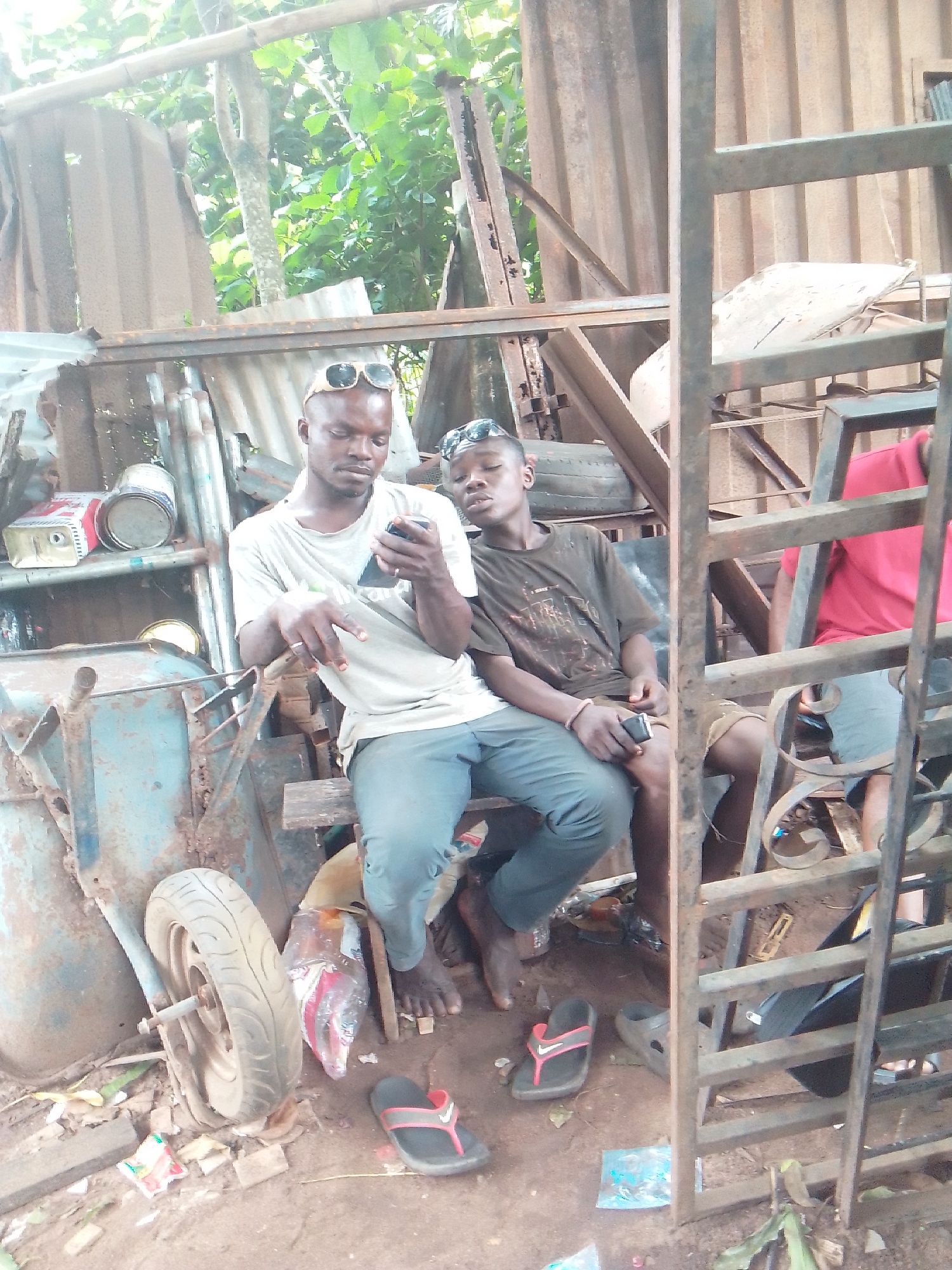 This image captures two males seated within a cluttered outdoor setting, likely a makeshift workshop or storage area. The man on the left, an adult, wears a light-colored T-shirt, dark trousers, and has sunglasses resting on his head. He is barefoot, with a pair of black and red flip-flops on the ground near him. He holds a black smartphone in his hands, focusing intently on its screen. A ring is visible on his left hand.
Beside him, a younger male wears a dark T-shirt with a faded graphic, shorts, and also has sunglasses on his head. He is barefoot, with dark flip-flops on the ground nearby, and holds a small black object in his left hand. He appears to be observing the adult or looking slightly past him. A third person, wearing a red T-shirt, is partially visible in the background on the right, behind a vertical metal frame.
The setting is characterized by a lean-to structure made from corrugated metal sheets and metal beams, with lush green foliage and trees visible in the background, suggesting a tropical or sub-tropical environment. To the left, a rusty blue wheelbarrow with a single wheel is visible, along with several metal cans, one with a red and white label partially visible, possibly indicating paint. The ground is bare earth, strewn with plastic wrappers and debris. The lighting indicates it is daytime. The scene conveys a relaxed, informal atmosphere.