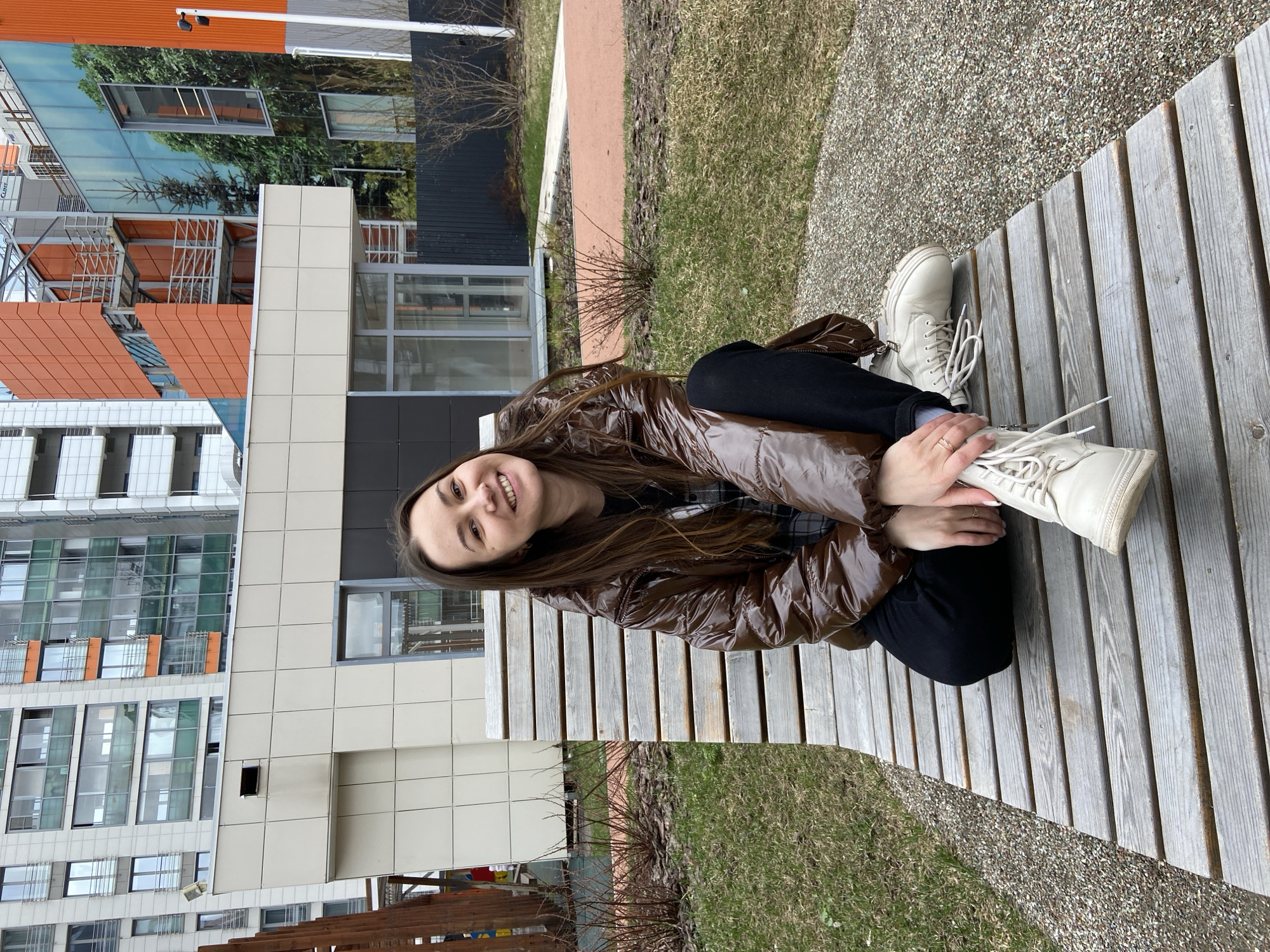 The image features a young woman smiling brightly while sitting on a wooden slatted bench in an outdoor urban environment. She has long, dark brown hair and is dressed in a shiny, puffy brown jacket, black trousers, and light cream-colored combat boots. A ring is visible on her finger as her hand rests on one of her boots.
The background showcases modern architecture, including multi-story buildings with a mix of light-colored facade panels and large glass windows. Some buildings feature distinct orange accents, and reflections of trees and the sky can be seen in the glass surfaces. The ground around the bench is covered with a gravel path and patches of sparse, yellowish-green grass, along with some bare shrubs and tree branches, suggesting an early spring or late autumn season. The lighting is soft and bright, indicating daytime, possibly under an overcast sky, as there are no harsh shadows. The woman appears happy and relaxed, seemingly posing for the camera. The scene is located in Izhevsk, Russia. No visible text is present in the image.