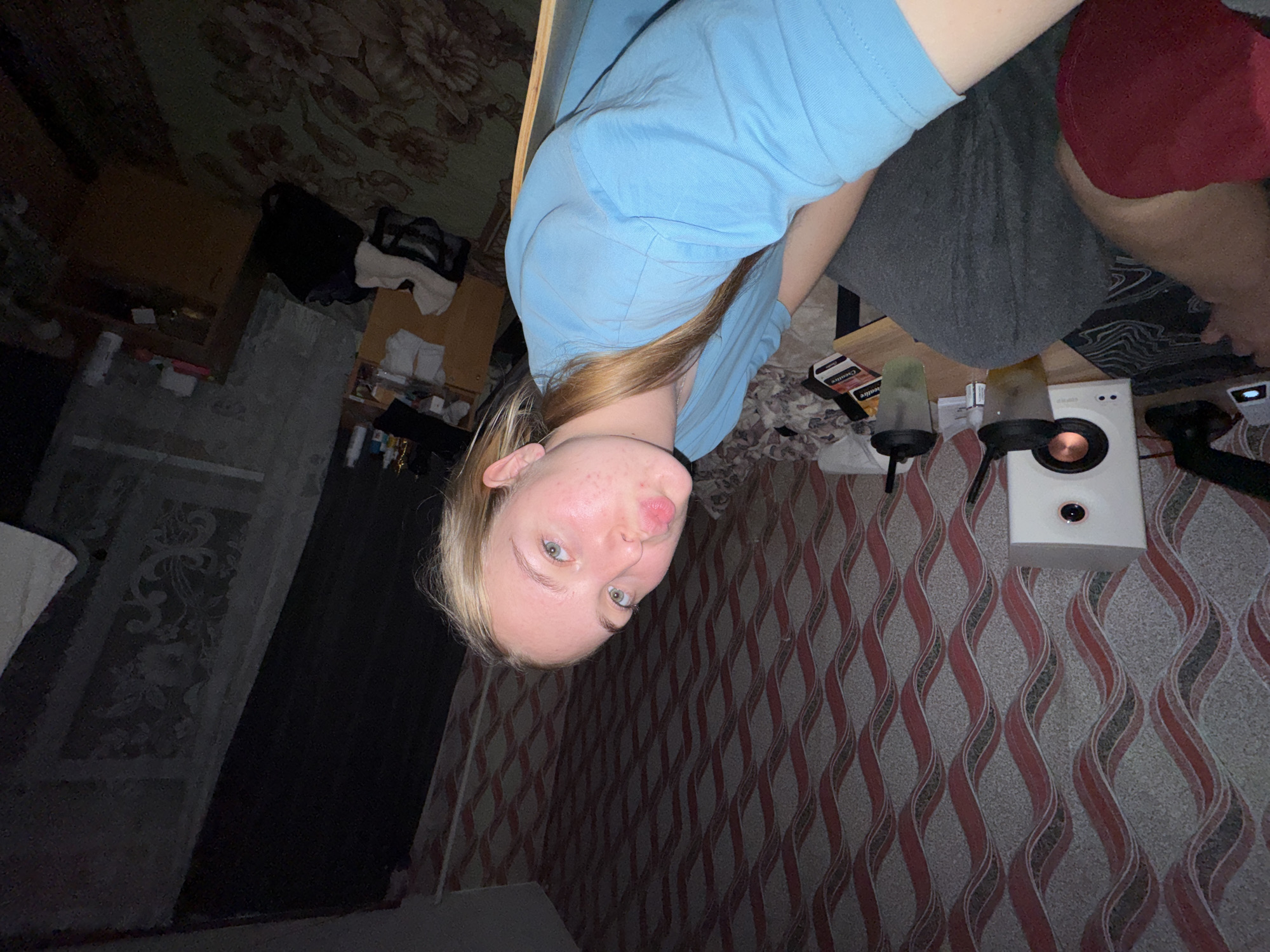 A medium shot, slightly rotated, shows a fair-skinned young woman with blonde hair partially covering her blue shirt. She is positioned in front of a patterned wallpaper with red and grey waves. Adjacent to her, on a table, are a white speaker, two microphone stands, and several small boxes. She sits at the table, partially visible in a grey garment. Another person, wearing red clothing, is seated beside her. The background displays parts of a room with floral print carpet, furniture, and personal items. The image was captured in Perm, Russia.
