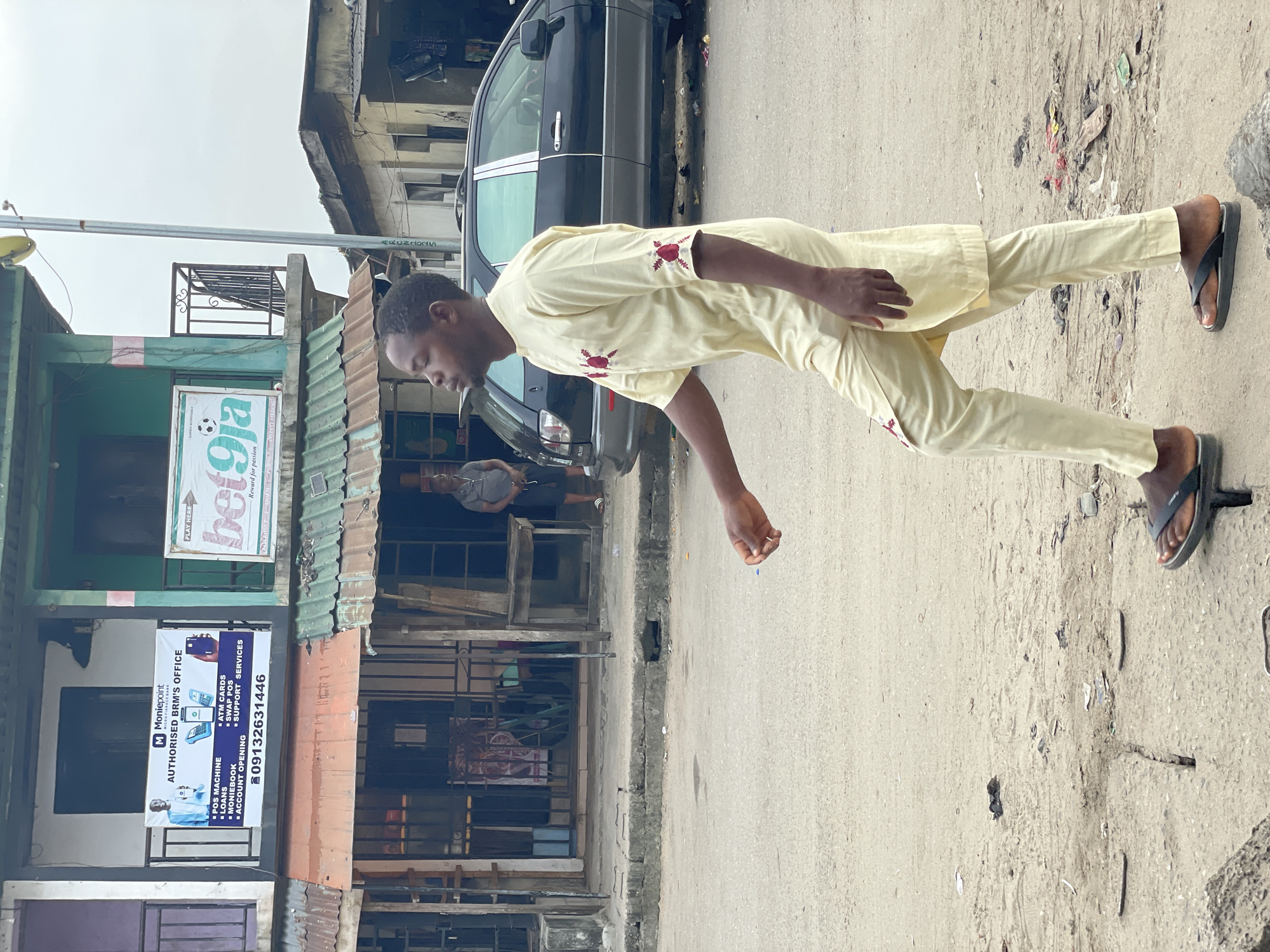 A man in a light yellow traditional outfit with red embroidery on the sleeves and hem walks along a dirt and asphalt road in Ado, Nigeria. He is wearing black flip-flops. In the background, a black SUV is parked on the side of the road. Behind the SUV is a building with multiple storefronts. One storefront displays a banner for "Moniepoint Authorized BRM's Office" with services listed such as POS Machine, Loans, Moniebook, Account Opening, ATM Cards, Swap POS, and Support Services. Another storefront has a sign that reads "bet9ja" with a soccer ball graphic. The environment appears to be an urban commercial area with weathered buildings and a dusty street. Other people are visible in the background near the buildings.