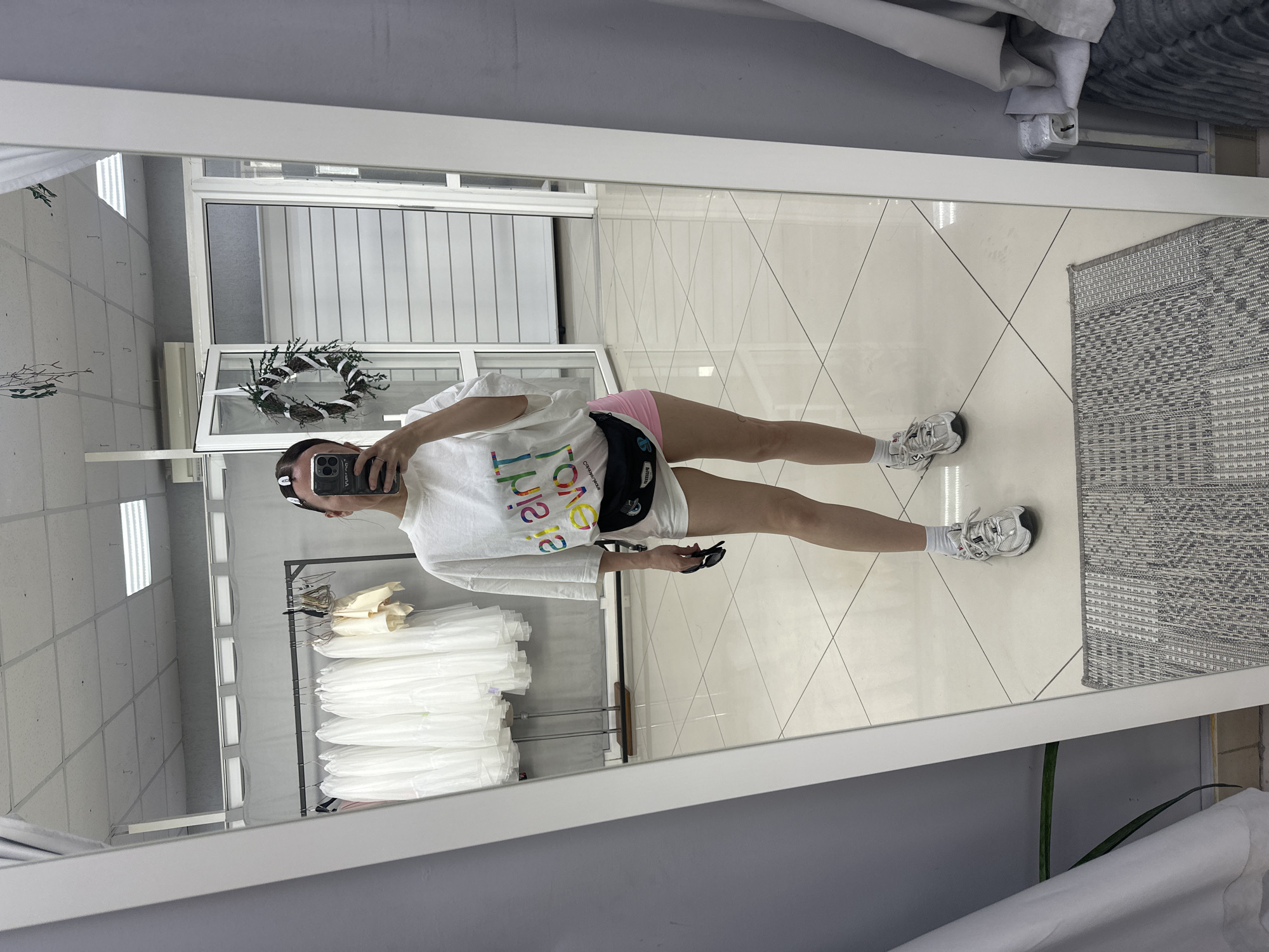 A person is taking a mirror selfie. The person is wearing a white t-shirt, pink shorts, white socks, and white sneakers. The person is holding a black object in their hand. The person is standing in a room with a white tile floor and a gray wall. There is a rug on the floor. There is a rack of white clothing in the background. There is a wreath hanging on the wall. The image does not include definitive location information or evidence for Votkinsk, Russia.