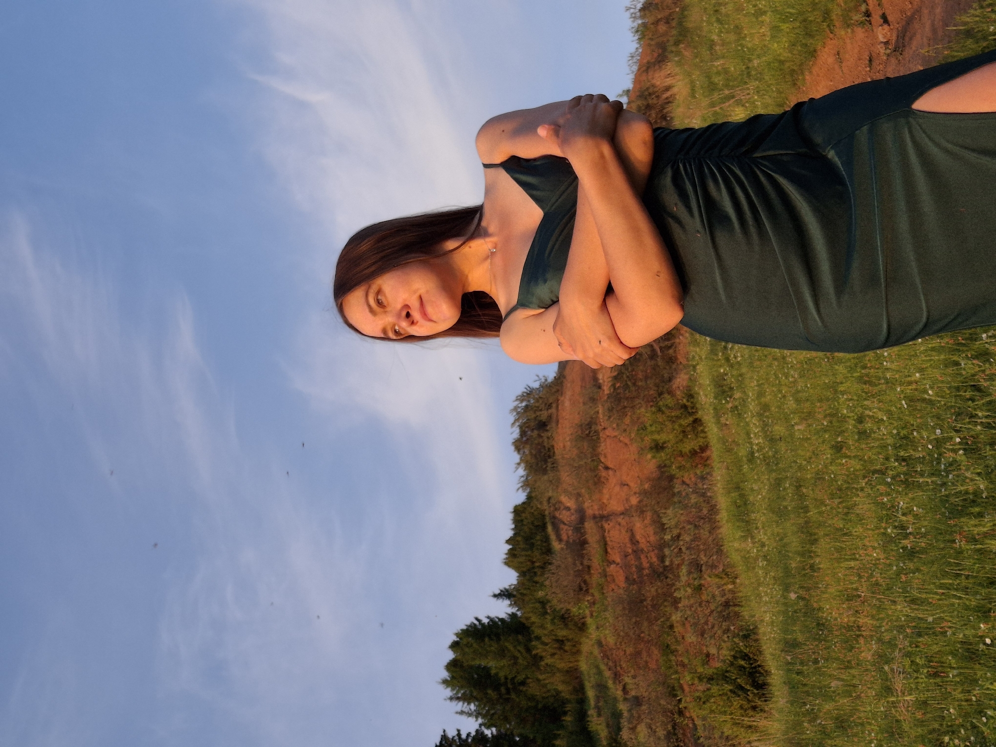 A woman is standing in a field in Ольховка, Russia. She is wearing a dark green dress with a slit on one side, and she has her arms crossed. The field is grassy and there is a dirt path running along the edge of it. Behind the field is a cliff with trees growing on top. The sky is blue with some white clouds.