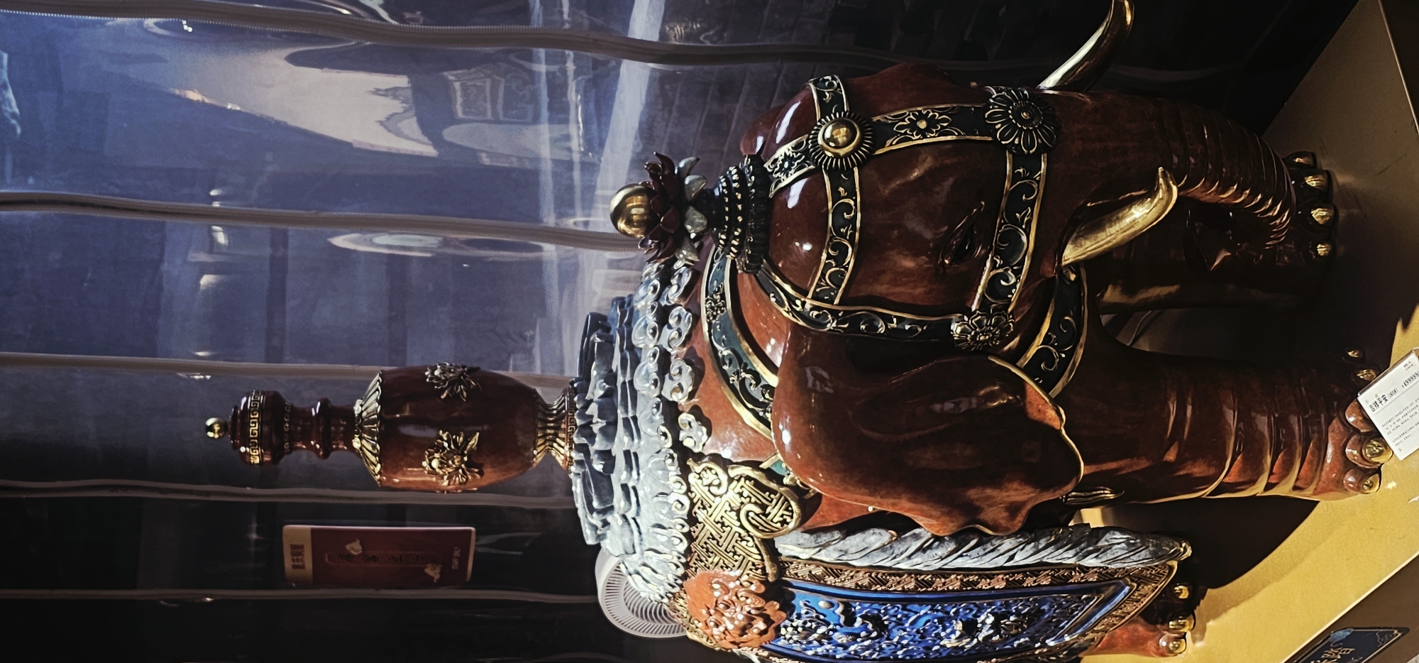 This is a close-up, vertical, eye-level shot of an ornate, reddish-brown ceramic elephant statue displayed indoors. The elephant is elaborately decorated with gold and blue accents, including intricate patterns on its blanket, harness, and headdress. A tall, dark red vase with gold embellishments sits atop the elephant's back, supported by a decorative base featuring stylized clouds. The background is blurred and consists of vertical lines, possibly representing curtains or a window, with reflections of an interior space and some outdoor lighting visible. A white fan is partially visible to the left of the elephant. A small white sticker with text is placed on the golden platform in front of the elephant. The image appears to be taken at an exhibit or a display within Yuzhong District, China, likely in a museum or gallery setting, given the presentation of the artifact. There are no people visible in the frame.