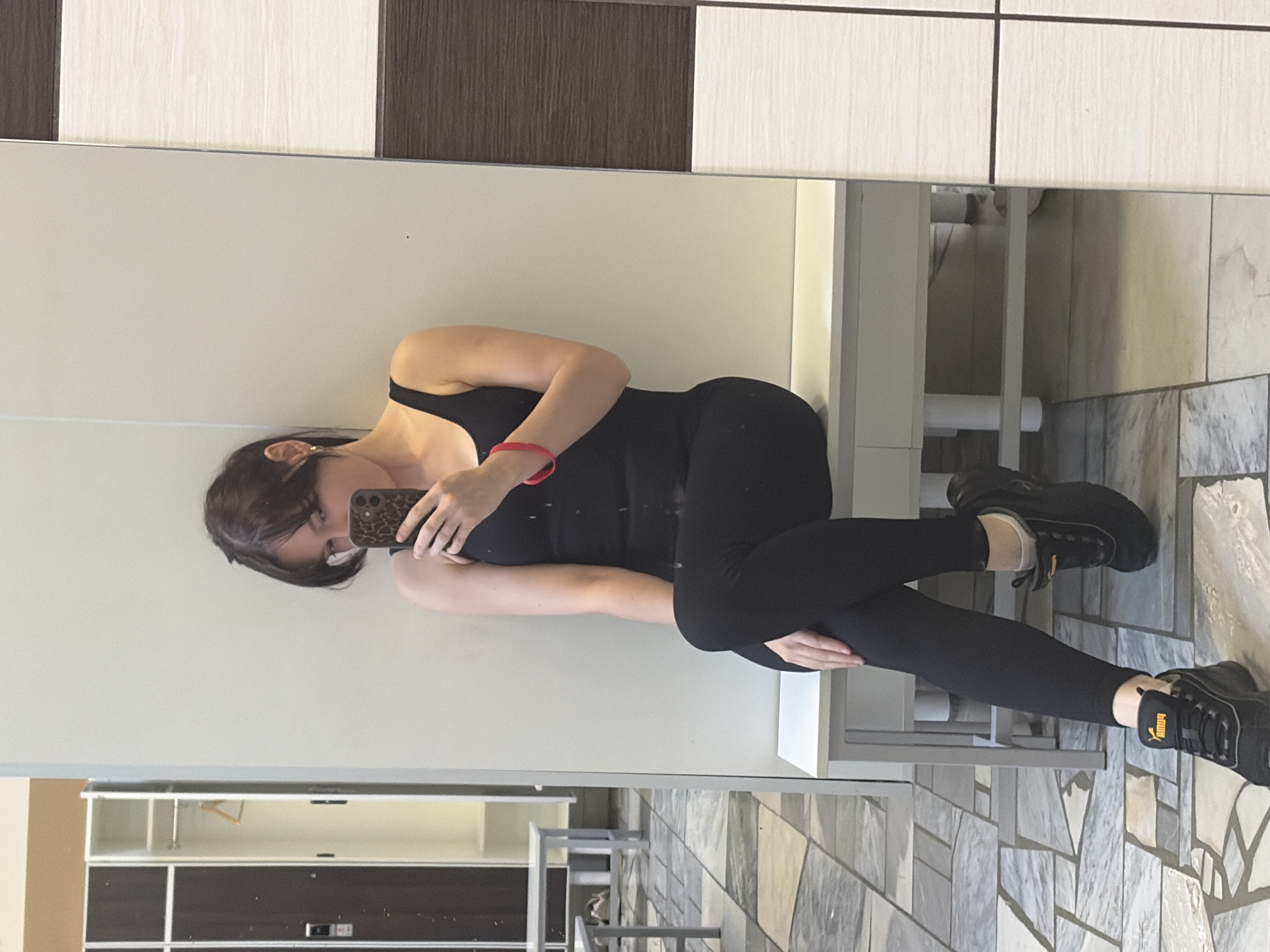 A young woman with dark hair and bangs, wearing a black tank top, black leggings, and black Puma sneakers, is taking a selfie in a locker room. She is sitting on a white bench and holding a phone with a leopard-print case. A red bracelet is visible on her left wrist. The locker room has tiled flooring, white walls, and dark brown and white lockers. The scene suggests a gym or sports facility. The context of Chaikovsky, Russia is not visually evident in the provided image.