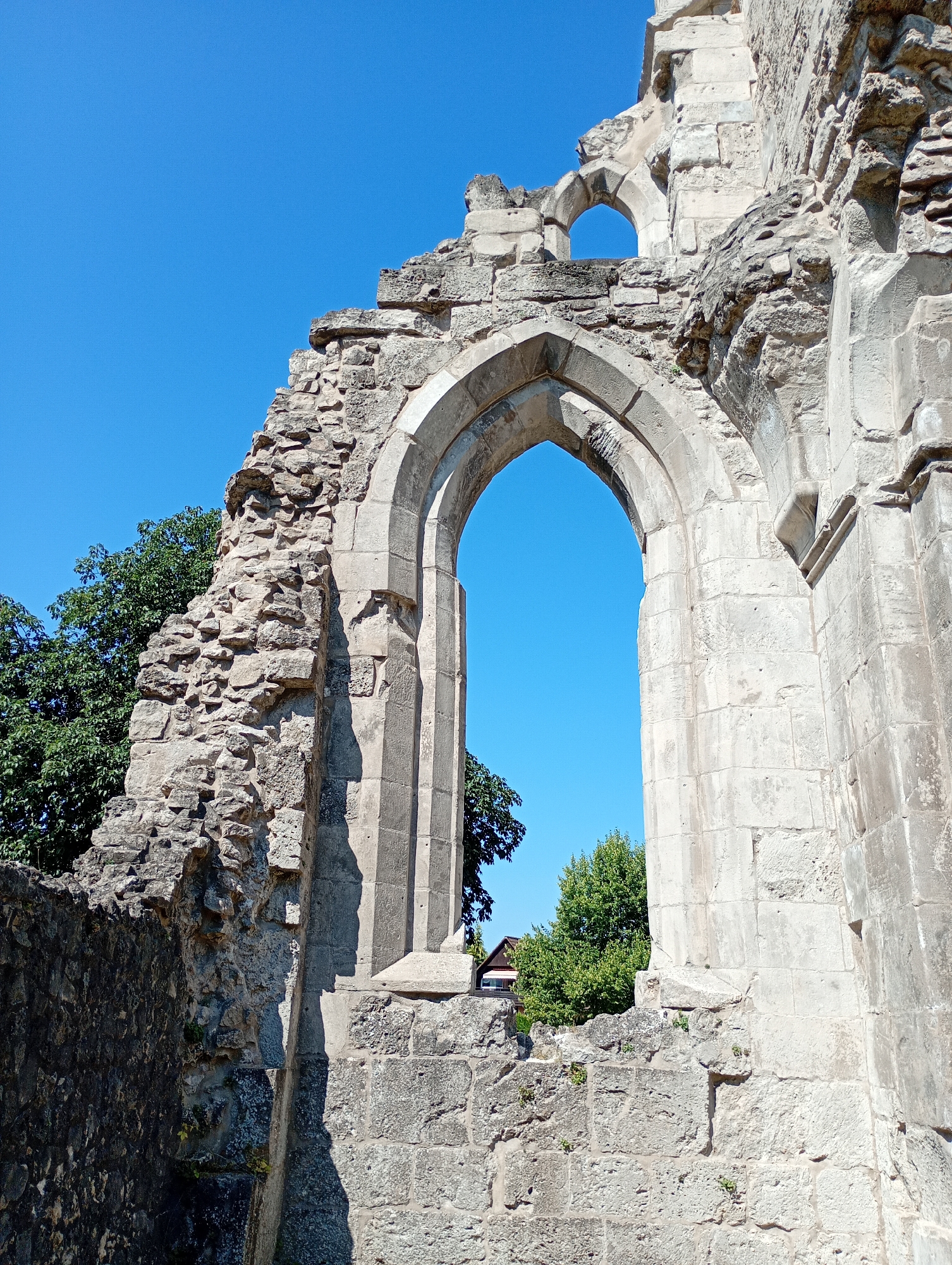 The image shows the ruins of a gothic arched window with a bright blue sky visible through it. The stonework is weathered and crumbling, with large, rough-hewn blocks forming the structure. Part of a green tree is visible on the left side of the frame, and another tree with a small building behind it can be seen through the archway. The ruins are located in the city of Walkenried, Germany, and appear to be part of an old church or abbey. The lighting suggests it is daytime. There are no people or animals visible in the image.