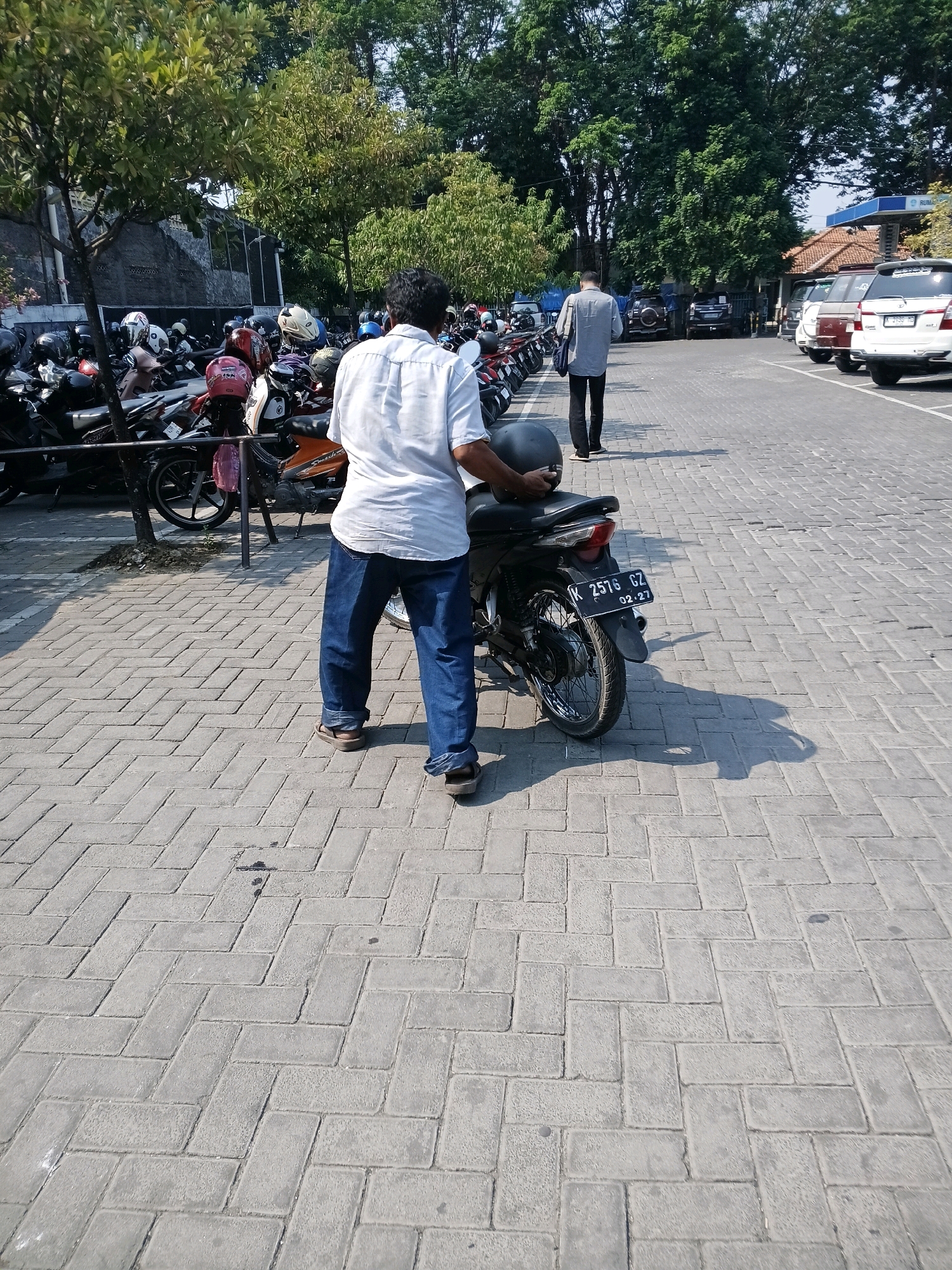A man stands facing away from the camera in a paved parking lot in Purwodadi, Indonesia. He is wearing a white short-sleeved shirt and dark blue jeans. In his right hand, he holds a black motorcycle helmet. Next to him is a black motorcycle, license plate K 2576 GZ. To the left of the man and motorcycle are many other parked motorcycles, some with helmets on them. To the right of the man and motorcycle, the parking lot continues, with a few cars parked in the distance. Trees line the background.