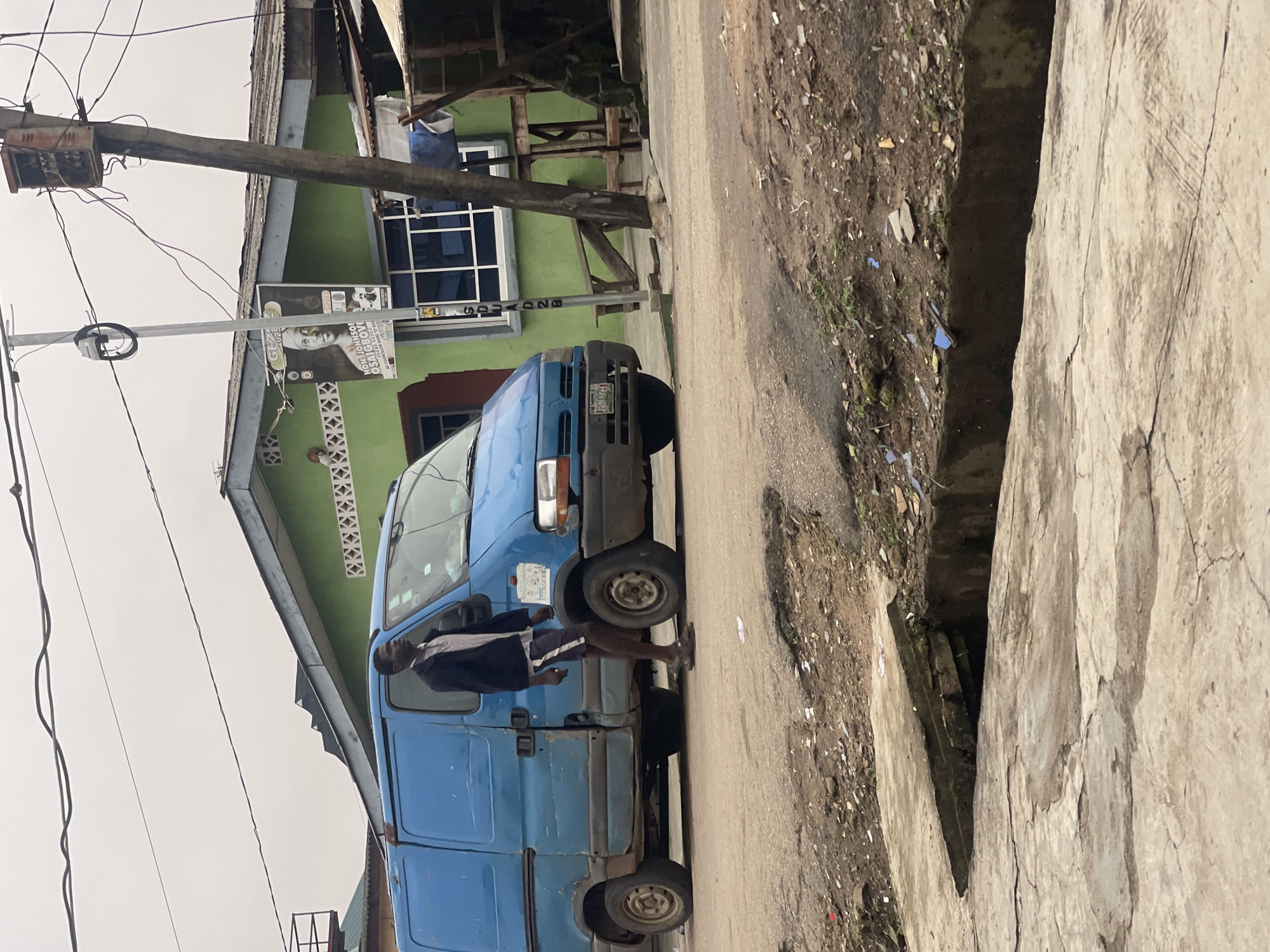 A man stands beside a light blue van in what appears to be a residential or commercial area in Akowonjo, Nigeria. The man, wearing a dark jacket over a light shirt and dark shorts with white stripes, stands to the left of the van, facing forward with his left hand in his pocket and his right arm slightly bent. The van is positioned on a paved road that shows signs of wear and tear, with debris and dirt scattered on the surface and along the sides.
Behind the van, a one-story building with a bright green exterior is visible. It has several windows, some with white frames and bars, and a dark-colored roof. A poster is attached to a pole in front of the building, featuring a portrait of a man and text that appears to be related to an election or political campaign, with the name "HON. JOHNSON OSAIGBOVO" and "COUNCILLOR WARD 13" legible. A utility pole with various wires and an electrical box stands to the right of the building.
In the foreground, a concrete drainage channel runs across the image, with a section of its cover dislodged and tilted. The surrounding ground is uneven, with patches of dirt, vegetation, and scattered debris. The sky is overcast, contributing to the muted lighting of the scene.