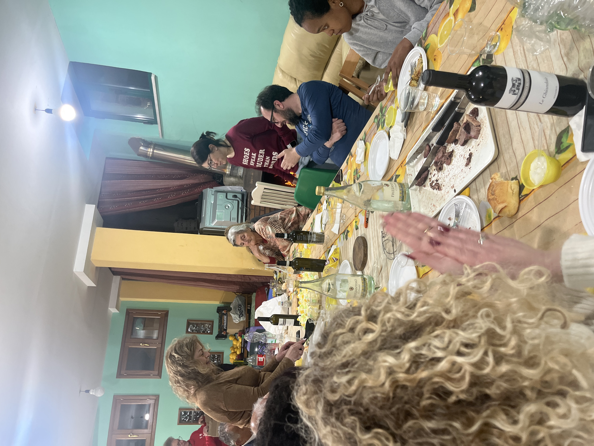 Here is a description of the image:
**Scene:**
The image depicts a gathering of people around a long, food-laden table, suggesting a meal or celebration. The event appears to be taking place indoors.
**People:**
There are multiple people in the image, including adults and what seems to be one child who is partially seen. The group is mixed in gender and ethnicity. People are sitting and engaged at the table, with some focused on eating and others involved in conversation.
**Objects:**
The table is covered with a lemon-themed tablecloth and filled with an assortment of food, including what looks like sliced meat, bread, and other dishes. There are plates, cutlery, glasses, and multiple bottles of wine on the table, one with a label that says "Le Chiandave". Other text indicates it's Frizzante wine. Decorations and personal items are also present, adding to the lived-in feel of the space.
**Actions and Interactions:**
People are eating, drinking, and socializing. The focus is on enjoying a meal together. Some individuals are actively serving food, while others are engaged in conversation.
**Environment:**
The setting includes a blend of old and new aesthetics. A fireplace area can be seen in the background, along with a window providing natural light. Various decorative items and artwork are visible in the background. The interior color scheme includes pastel green and yellow.
**Location Notes:**
Bottled water and wine with the words "Vingiani" and "Frizzantino" along with "Alfonso Rotolo" and "Le Chiandare" suggest that the location may be Cannalonga, Italy, given the presence of wineries there bearing the Rotolo name.