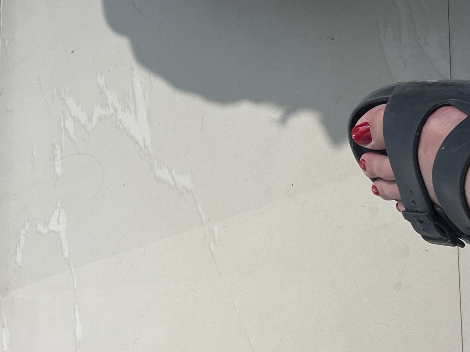 A close-up, low-angle, eye-level shot shows a person's left foot wearing a dark grey sandal with two straps. The toes are painted with bright red nail polish. The sandal is positioned on a light-colored, tiled floor with faint water stains and a thin, dark line across the floor, possibly a grout line. A soft shadow, cast from a person, is visible on the floor to the right of the foot and sandal, partially obscuring the floor pattern. The lighting suggests an indoor setting with natural light source from the right. The image is focused on the foot and sandal, with the floor and shadow as background elements. The location is unknown.