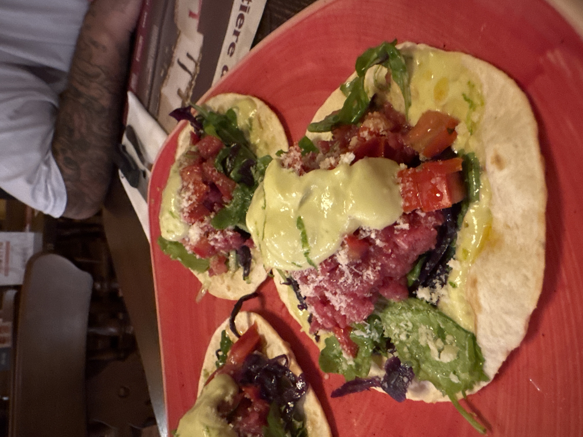 Three tacos are presented on a red platter. The tacos have a thin, circular tortilla base topped with finely chopped red meat, diced tomatoes, greens, and a light green sauce, possibly avocado-based. Grated white cheese is sprinkled over the fillings. In the background, a person's arm with tattoos is visible, resting on a table. A fork and knife are also present on a white napkin. The setting appears to be an indoor restaurant.