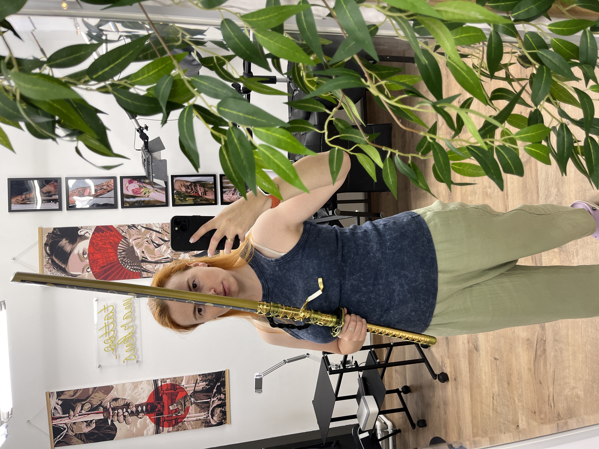 In a room with a white wall, a woman with red hair, wearing a dark blue tank top and light green pants, stands facing a mirror and taking a picture with her phone. She is holding a decorative sword with a golden hilt and scabbard, with the blade positioned vertically in front of her face, obscuring her eyes and nose. The room appears to be a tattoo studio, evidenced by the various tattoo-themed artwork displayed on the walls, including a large illustration of a geisha with a red fan and another featuring a samurai. A neon sign with yellow lettering, possibly "Tattoo Studio," is also visible. In the background, there is tattoo equipment, including a black tattoo chair and a rolling table with a device on it. A branch with green leaves hangs in the foreground, partially obscuring the reflection in the mirror. The floor is made of wooden planks. The scene is well-lit, suggesting indoor lighting. No specific location in Chaikovsky, Russia can be determined from the image.