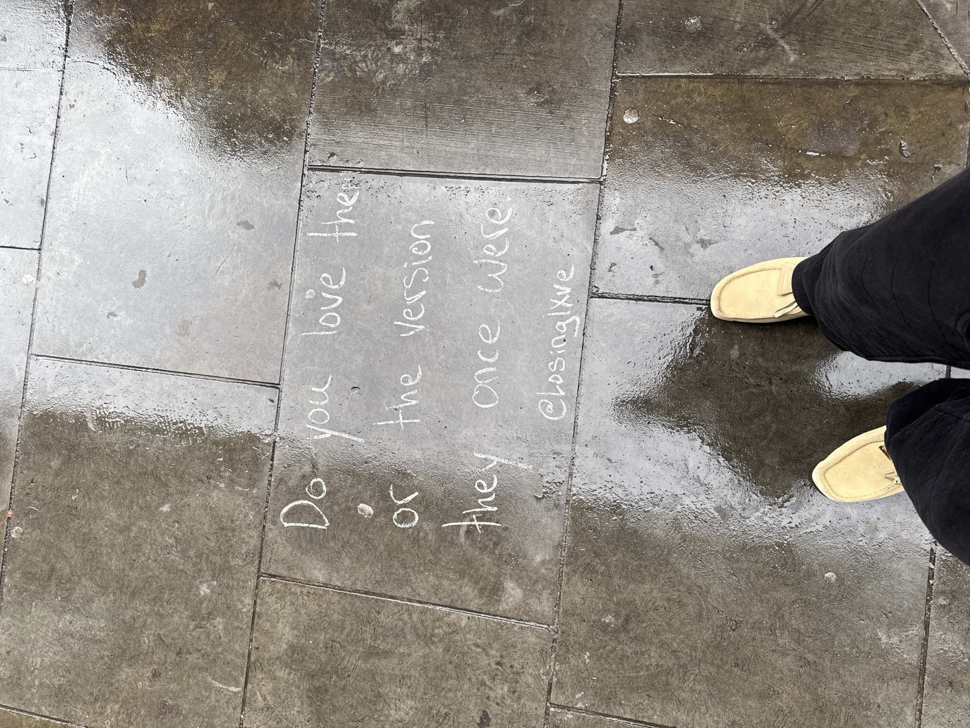 The photograph shows wet pavement in an urban setting with white chalk writing that reads "Do you love the or the version they once were. @closinglxve". To the right of the writing are the feet of a person wearing black pants and tan loafers. The scene is situated on a sidewalk paved with rectangular concrete slabs, reflecting the surrounding environment due to the wet surface. Based on the Instagram handle visible (@closinglxve), it is highly likely this scene is located in the city of London, United Kingdom.