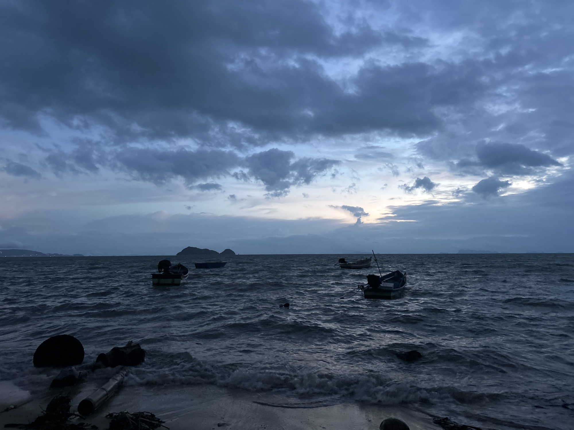 The image depicts a dark, cloudy sky over a choppy sea with several small fishing boats anchored offshore. The scene appears to be a beach, with debris like logs and plastic wash ashore in the foreground. The overall lighting suggests it is either dawn or dusk. The location is likely a coastal area in Thailand, possibly Ko Pha-ngan District, given the style of boats and the tropical landscape visible on the horizon. There are no people visible in the image.