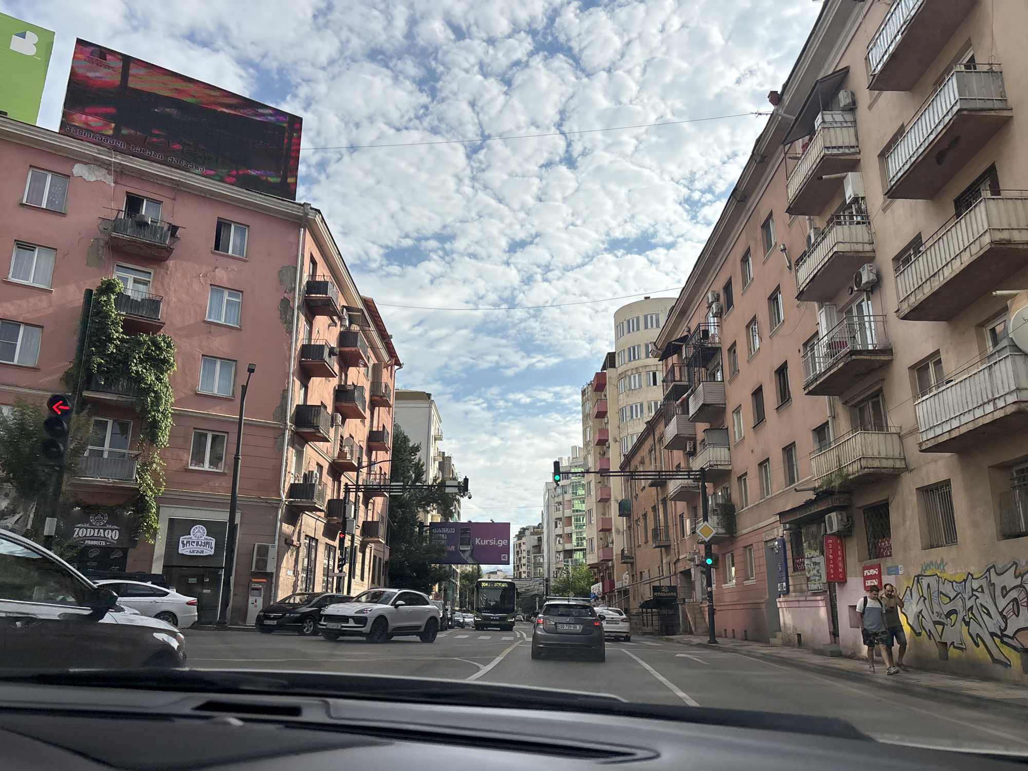 The image captures a street scene in Tbilisi, Georgia, likely on a day with a partly cloudy sky. The perspective is from inside a vehicle, looking forward. On the left, a pink apartment building features numerous balconies, some with air conditioning units. A traffic light with a red left-turn arrow is visible. Further down the street, a sign for "ZODIACO PRODUCTS" is present. On the right, another pink apartment building, also with multiple balconies, lines the street. Graffiti adorns a section of this building's lower facade. Two men are walking on the sidewalk, one wearing a backpack. Several cars are on the road, including a white SUV and a black sedan. A green bus with "304" displayed on its front is in the middle distance. In the background, other buildings of varying heights and architectural styles are visible, along with streetlights and utility wires crisscrossing the sky. A large billboard with an advertisement is mounted high on the left side.