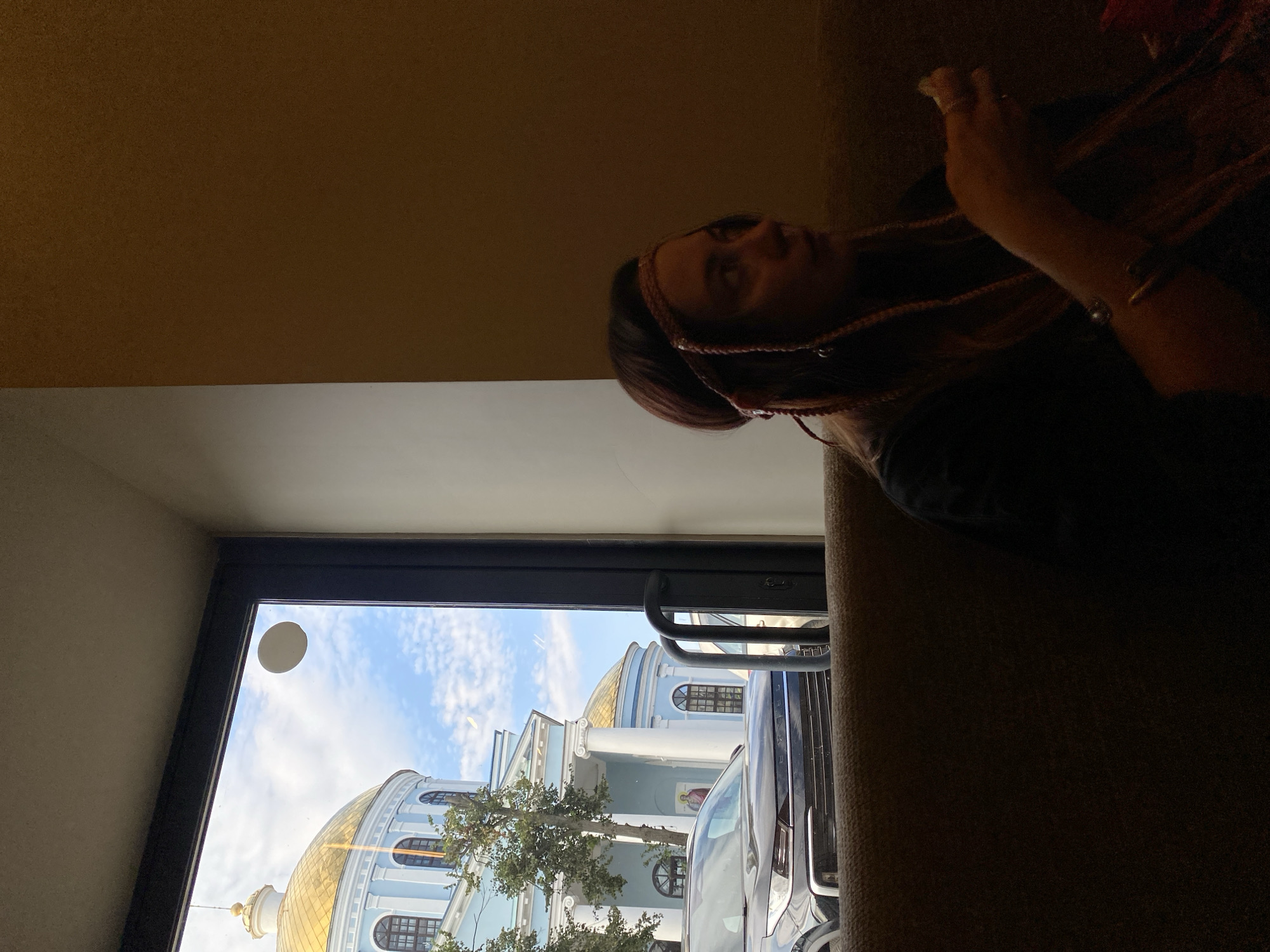 The image captures an indoor scene looking out a large window. A woman with braided hair and a decorative headband is seated on a brown couch, partially visible in the foreground. She appears to be eating. Through the window, a light blue building with a golden dome and classical architectural features, resembling a church or government building, is visible under a partly cloudy sky. A dark-colored SUV is parked in front of the building. The overall setting suggests a cafe or restaurant with a view of a prominent landmark in the city of Votkinsk, Russia.
