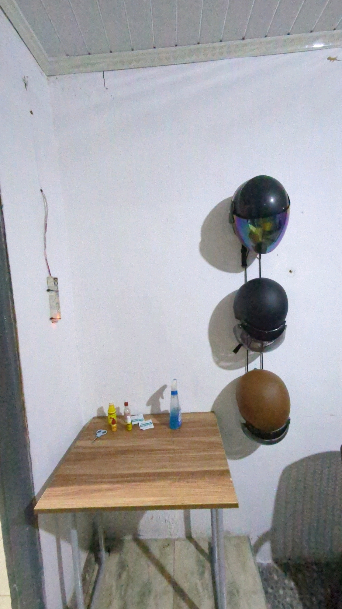 The image depicts an indoor scene, likely a corner of a room. A light-colored ceiling with parallel paneling is visible at the top. The walls are white and textured, with some minor cracks and a dangling electrical wire with a small device attached.
In the corner, a small, light-brown wooden table with metal legs stands on a gray tiled floor. On the table, there are a few small items: a pair of scissors, two small bottles (one yellow, one red), and a blue spray bottle.
To the right of the table, mounted on the wall, is a rack holding three motorcycle helmets. The top helmet is black with a iridescent visor, the middle helmet is black and round, and the bottom helmet is a brown, egg-shaped open-face helmet. The helmets cast distinct shadows on the wall.
The image does not contain any specific elements that would allow for pinpointing its location to Baroqah, Indonesia. The scene is a generic interior setting.