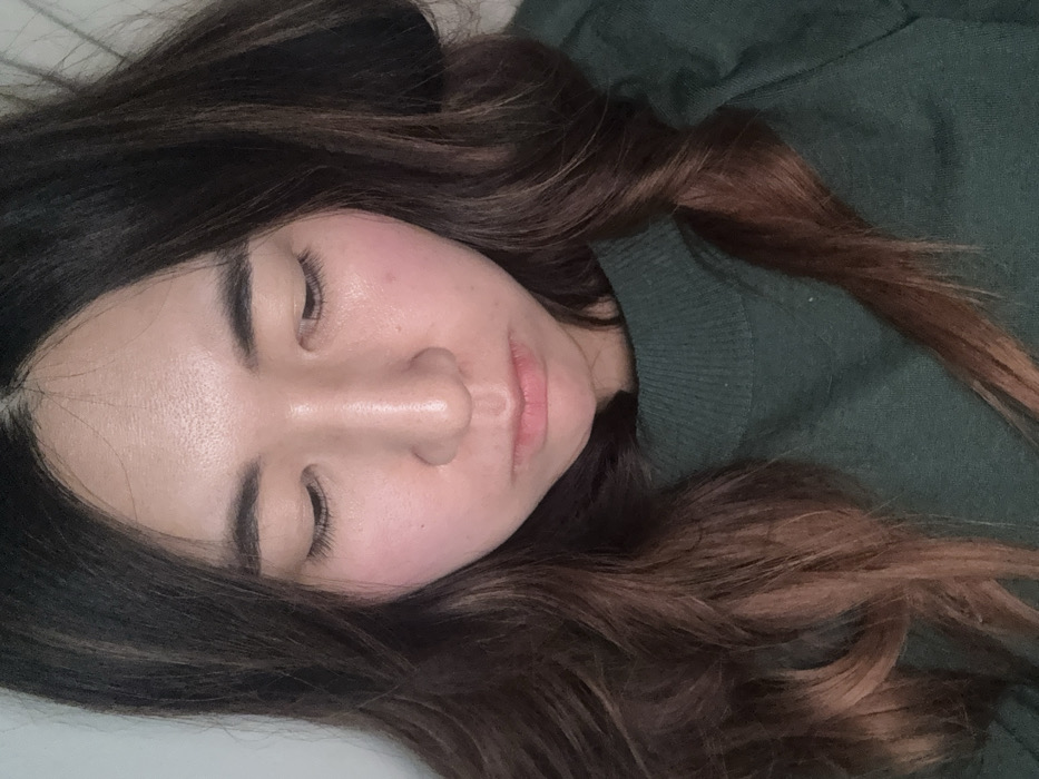 The scene is an indoor setting in Paris, France. A young East Asian female is depicted in a close-up, top-down perspective, looking downwards. Her eyes are cast towards an unseen point, and her lips form a subtle downturn. She has long, wavy dark brown hair with lighter, possibly auburn, highlights, parted centrally and framing her face. Her complexion is fair with visible rosy cheeks. She wears a dark green, ribbed, high-neck sweater. The immediate background behind her appears to be a plain, light-colored, indistinct surface.