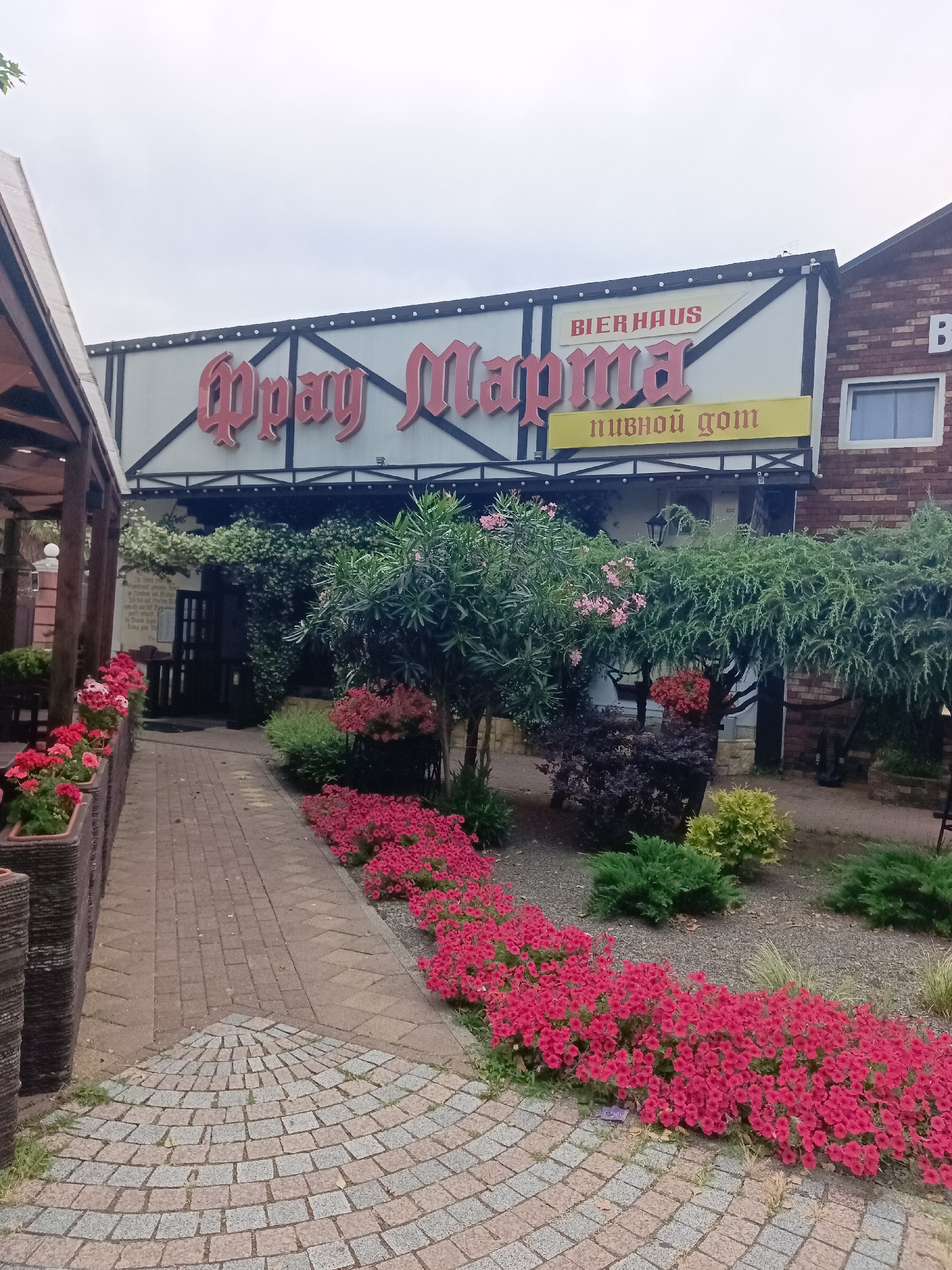 The image shows the facade of a building with a sign that reads "Bierhaus" and Cyrillic text that translates to "Beer House." The building has a traditional Bavarian architectural style, with dark timber accents and white stucco walls. It is surrounded by a garden with lush greenery and vibrant pink flowers. A paved walkway leads to the entrance, with potted plants and decorative elements enhancing the ambiance. The scene suggests a beer garden or restaurant, possibly a tourist attraction in Sochi, Russia, known for its coastal charm and recreational activities. The sky is overcast, indicating a cloudy day.