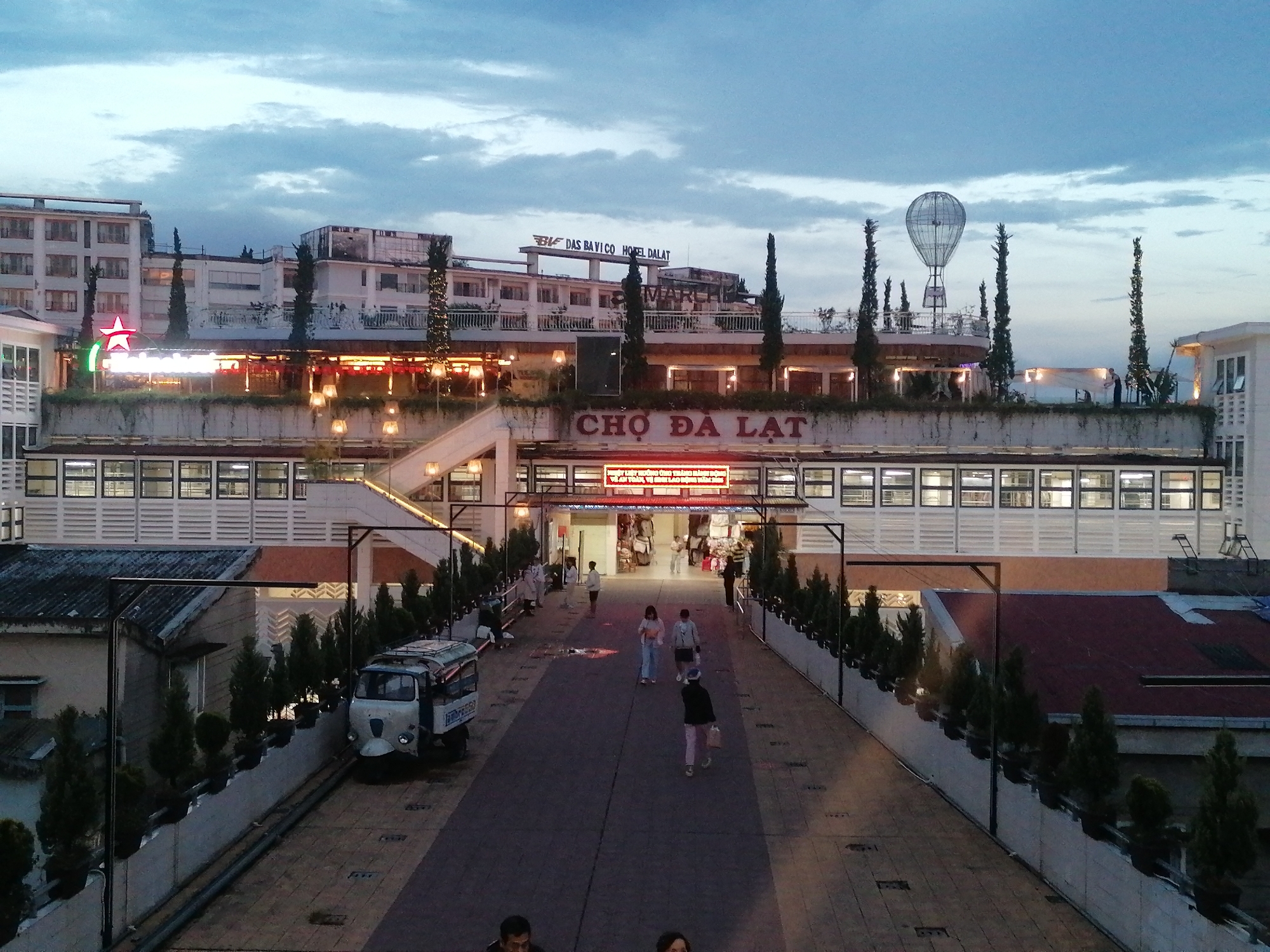 The image shows a slightly elevated view of Cho Da Lat, the central market in Da Lat, Vietnam, during the early evening or late afternoon. In the foreground, a paved pedestrian walkway stretches towards the market entrance, with rows of small evergreen trees in raised planters lining both sides. Several people are walking along the walkway, appearing as small figures. A white three-wheeled vehicle is parked on the left side of the walkway. The market building itself is a two-story structure with numerous windows and a large sign in Vietnamese script over the entrance. Above the market, several multi-story buildings are visible. On top of one of them, there is a replica of a hot air balloon. The sky is cloudy and dimly lit, suggesting the time of day.