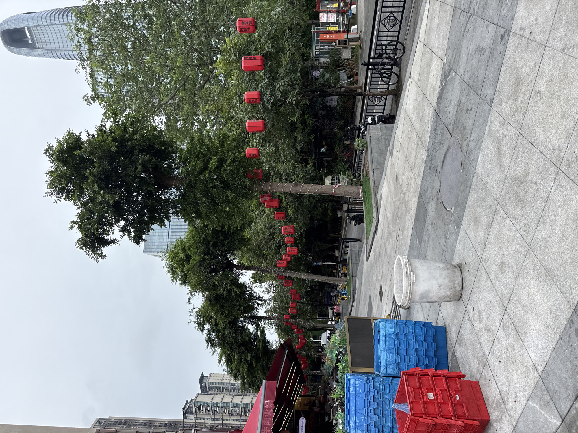 The image depicts an outdoor scene in Haizhu District, China, with a focus on a street lined with trees and urban architecture. In the foreground, several blue and red plastic crates are stacked on a tiled sidewalk, alongside a weathered white bucket. A red awning extends from a building to the left, suggesting a commercial establishment such as a restaurant. Red lanterns are strung between trees, adding a festive or decorative element to the environment. Beyond the immediate foreground, the street continues with more trees, a metal fence, a bicycle, and a parked scooter. In the background, tall modern buildings, including a distinctive skyscraper with a curved top, dominate the skyline under an overcast sky. The overall impression is of a bustling urban street scene, possibly in a commercial or entertainment district, with everyday objects and decorative elements coexisting with the impressive modern architecture.