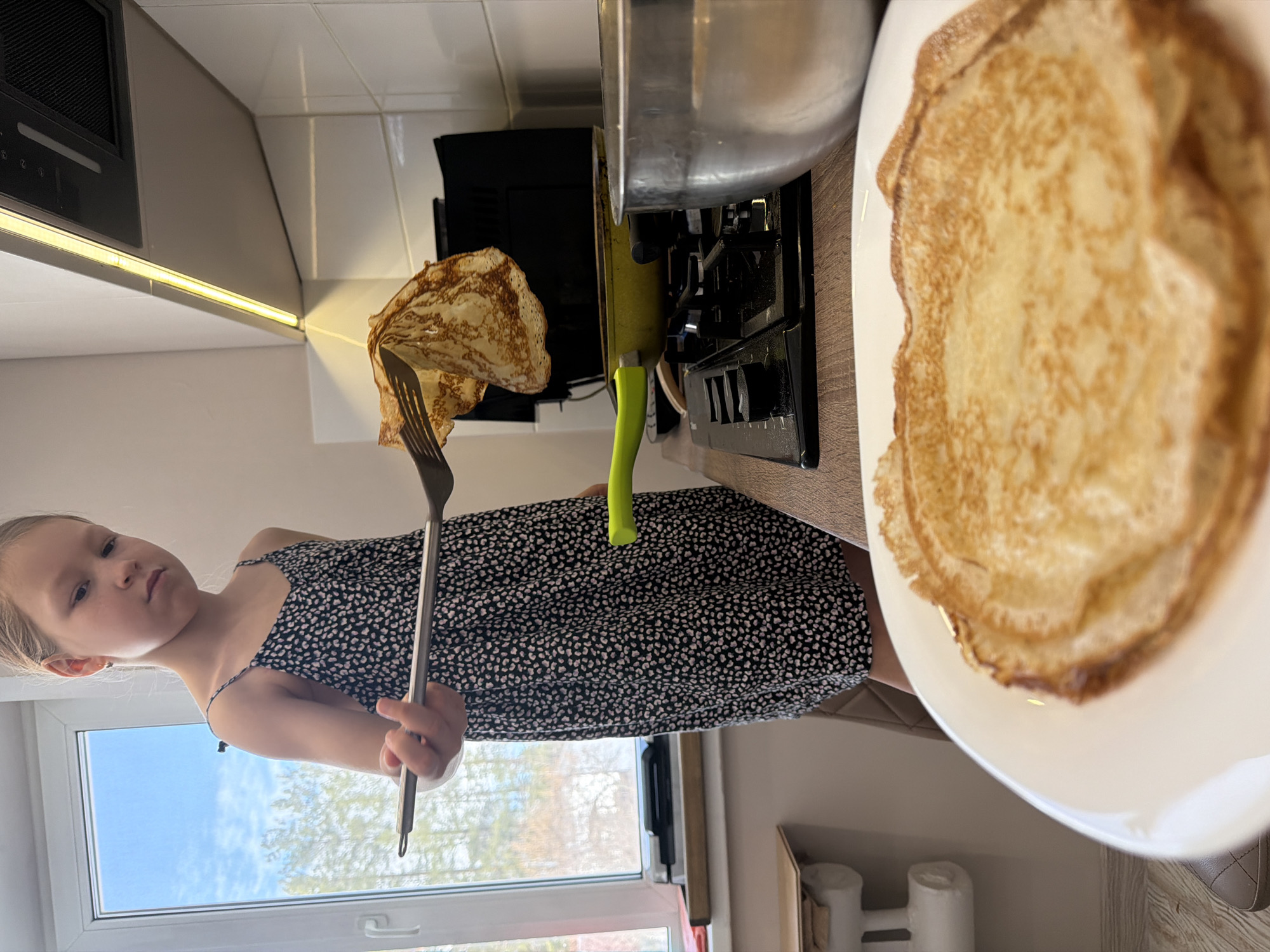 A young female child with light hair, wearing a black and white patterned spaghetti-strap dress, stands in a modern kitchen. She holds long metal tongs, using them to lift a golden-brown pancake over a yellow frying pan situated on a black gas stovetop. A stack of freshly cooked pancakes rests on a white plate in the immediate foreground. To the right of the stovetop, a large silver metal mixing bowl is present. The kitchen features white tiled walls and a light-colored countertop, illuminated by an under-cabinet LED light strip and natural light from a large window on the left. Through the window, a blue sky with white clouds and blurred green trees are visible. The location is a kitchen within the city of Unknown, Unknown.