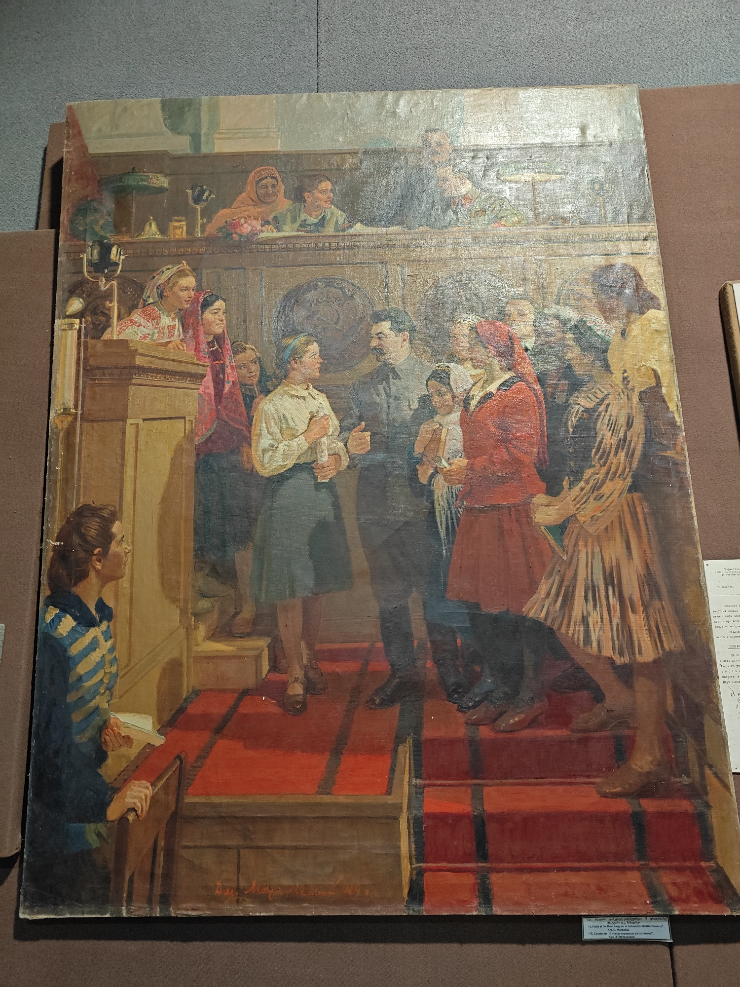 This painting depicts a formal indoor gathering, with a prominent male figure resembling Joseph Stalin at its center. He stands on a raised, red-carpeted platform, surrounded by several young women and girls. One woman, dressed in a white blouse and dark skirt, holds a rolled document, seemingly interacting directly with the central figure. Another woman in a red outfit holds a baby or small child. Many of the women are clad in colorful attire, some featuring headscarves and traditional-looking embroidered blouses.
To the left, a young woman in a striped blue and white shirt is seated at a wooden podium, observing the scene intently. The background shows more people, both on the main floor and in an upper wooden balcony, suggesting a large audience. The balcony also features decorative elements like a lamp, a bell, and a globe. The overall setting appears to be a grand assembly or meeting hall, characterized by ornate wooden paneling and structures.
The painting captures a moment of official interaction, possibly a presentation, reception, or congratulatory event. The mood seems formal and significant.
Visible text on a label below the painting, partly in English and partly in Russian, reads: "Stalin at the 12-nd congress of Advanced collective farmers" and "Art. D. M." followed by an illegible surname. Other Russian words like "Москва" (Moscow), "Предлож" (Proposal/Suggestion), and partially visible "ВОСТА" are also present, further contextualizing the artwork as a Soviet-era historical depiction.