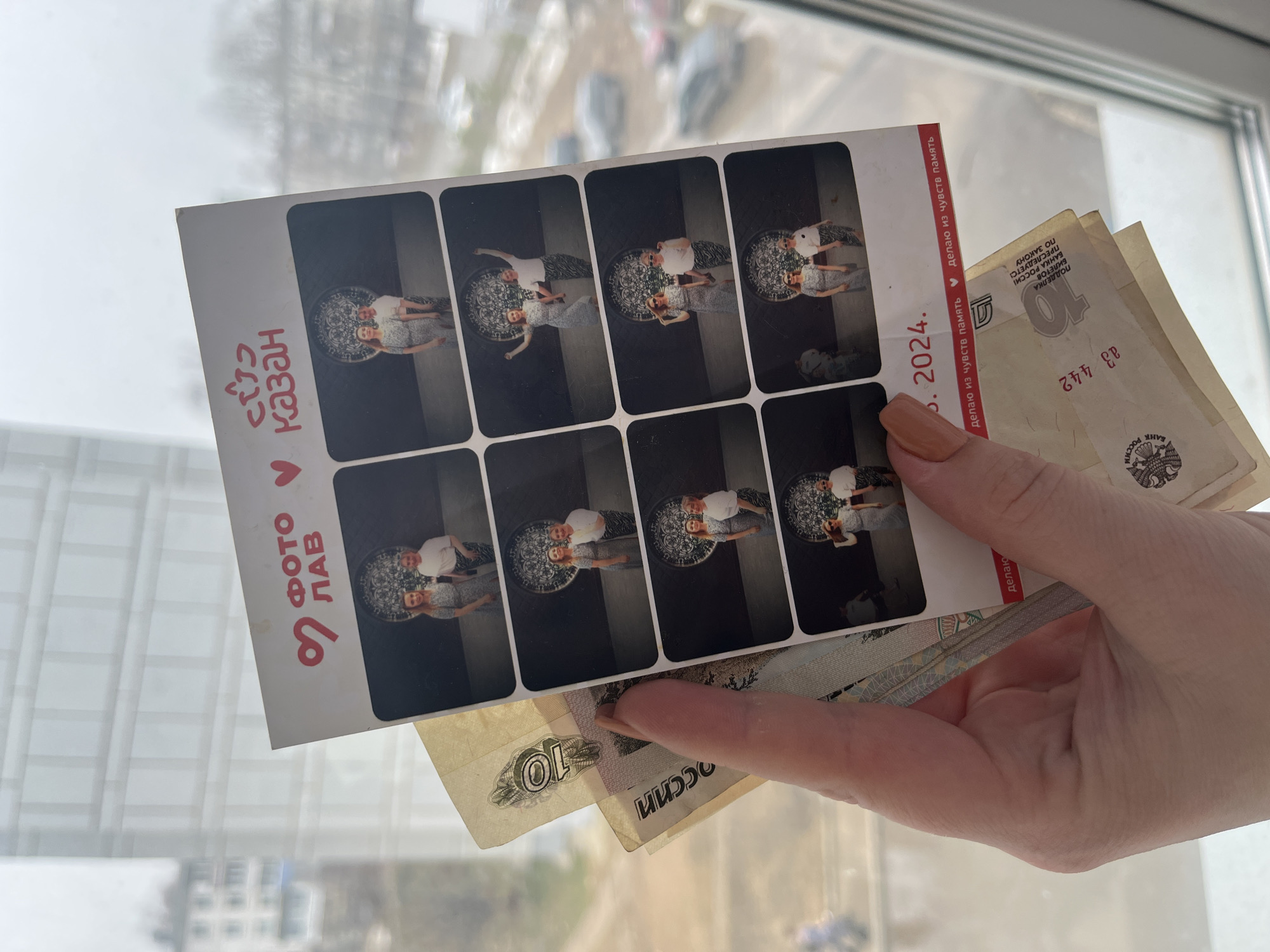 A photograph depicts a hand holding a strip of photo booth pictures and Russian banknotes. The photo booth strip contains eight identical images of a couple standing in what appears to be an interior with a patterned floor. The print on the side of the photo strip indicates "KAZAN." The banknotes in the hand are Russian rubles, with at least one visible as a 10-ruble note. The background shows a window and an outdoor urban scene. There is no evidence this image was taken in the city of Chaikovsky.
