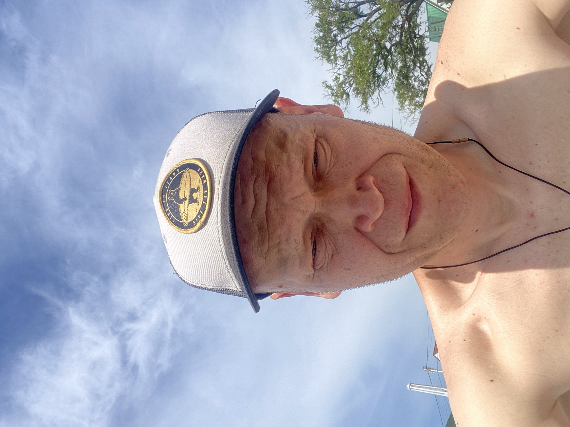 An adult male is depicted in a close-up, upward-angled shot under a clear blue sky with scattered wispy clouds. The subject is bare-chested, facing the viewer and looking downwards, with his eyes partially closed or squinting due to bright overhead sunlight. He wears a light-colored trucker-style baseball cap with a dark brim and a circular patch. The patch, dark with gold text and imagery, displays "LIFE IS JO___S" (the full word is obscured, but "JO" is clear) and "RIDE THE WAVE," featuring a central image of a bear on a surfboard. A thin, dark cord, possibly a necklace or earphone cable, hangs around his neck and over his right shoulder. Strong sunlight casts a distinct shadow from his head and cap onto his right shoulder. In the background, to the left, a tall, light-colored vertical structure, possibly a communication mast or pole, is visible. To the right, a portion of a green-leafed tree and what appears to be a building with a light-colored wall and a darker roof can be seen. The scene is located in Myskhako, Russia.