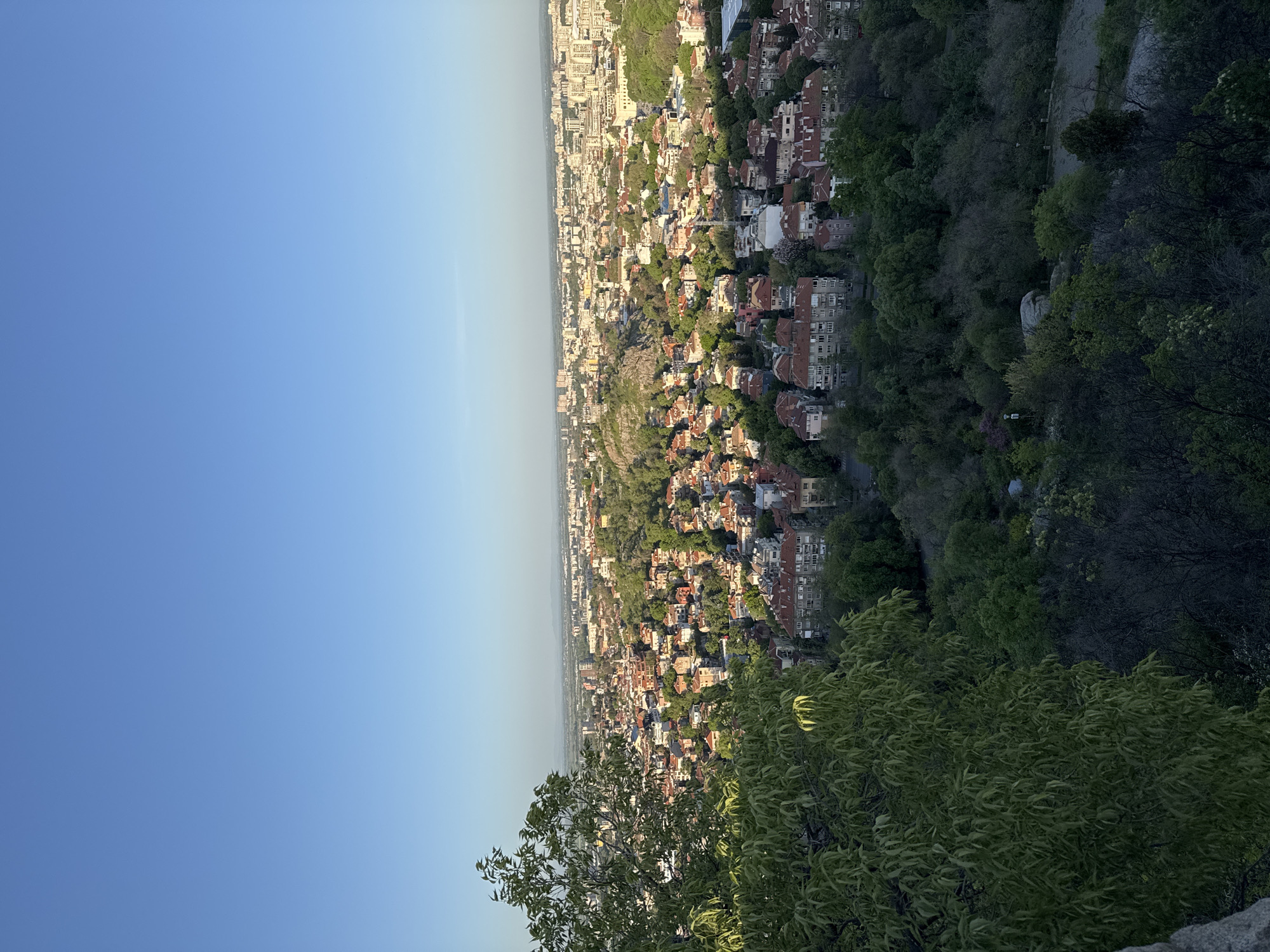This media file presents an elevated panoramic view of a city, likely Plovdiv, Bulgaria, from a vantage point offering a wide perspective of the urban landscape intertwined with natural elements. The image captures a vibrant cityscape under a clear blue sky, suggesting either early morning or late afternoon due to the angle of sunlight.
The scene is dominated by a densely packed residential area, featuring numerous buildings with predominantly red or orange tiled roofs. The architecture varies, with some areas showing older, more traditional structures and others appearing to be more modern multi-story apartments. The city stretches into the distance, with hazy contours of further urban development or distant hills visible on the horizon.
Interspersed throughout the urban sprawl and particularly prominent in the foreground and along the lower parts of the view are extensive green areas. Lush trees and foliage cover hillsides and natural rock formations, creating a striking contrast between the built environment and nature. Some trees are brightly illuminated by direct sunlight, while others are cast in deeper shadows, enhancing the perception of depth and the golden hour lighting. A large, lighter-colored rocky outcrop is visible mid-frame on the right.
The sky is uniformly clear and blue, fading slightly to a lighter hue towards the horizon, indicative of good weather conditions. There is no specific activity taking place, nor are there any people visible. No discernible text can be found within the image. The overall impression is one of a sprawling, historic city nestled within a green, hilly landscape.