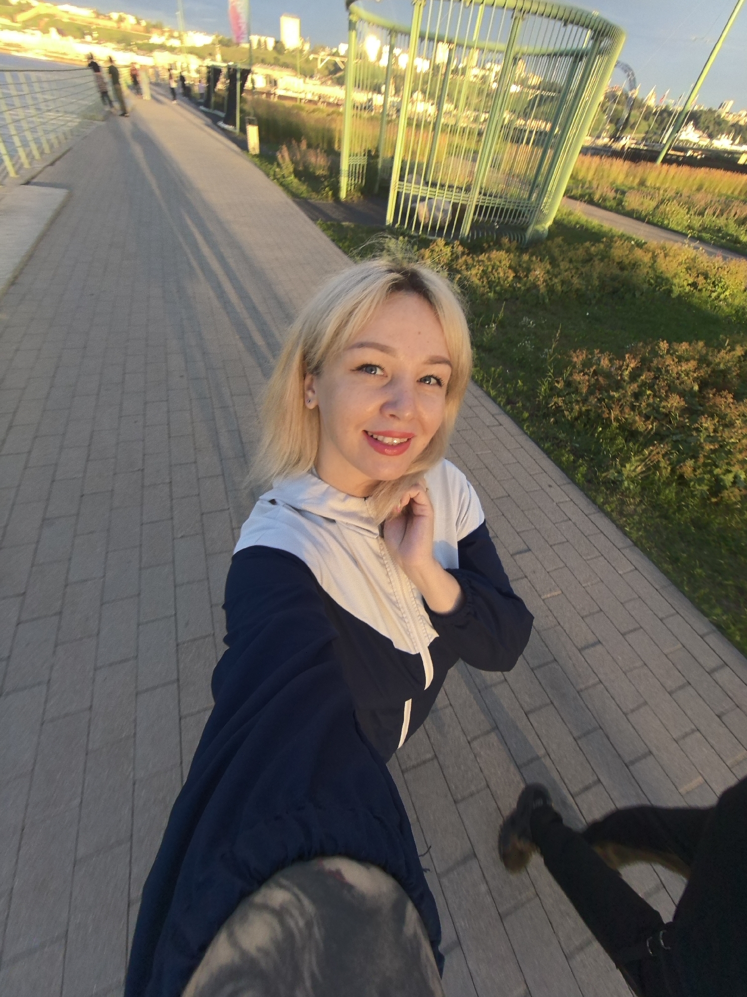 The image depicts a young woman with blonde hair, wearing a blue and white jacket, taking a selfie outdoors in Nizhny Novgorod, Russia. She is standing on a paved walkway that appears to be part of a promenade or park area. In the background, there is a distinctive green, cage-like structure and a body of water, likely the Volga River, with a city skyline and buildings visible across the water. The setting suggests a sunny day, possibly late afternoon, with long shadows cast on the walkway. Other people and what appears to be a dog are also present in the scene, though they are less prominent than the woman.