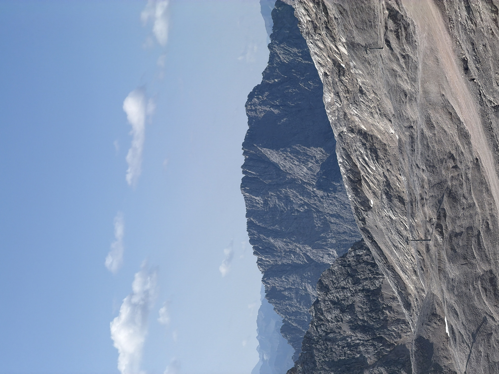 This is a scenic, high-angle, outdoor, daytime photograph of a mountainous landscape. The sky is a pale blue with scattered white clouds. In the foreground and midground, there are rocky, barren mountain slopes with a fine gravel surface, interspersed with patches of rock and soil. Several dark, slender poles, likely part of a ski lift or similar infrastructure, are visible on the slopes. The background features several layers of rugged, blue-grey mountain ranges, with the furthest mountains appearing hazy and distant. The scene depicts a natural, alpine environment.