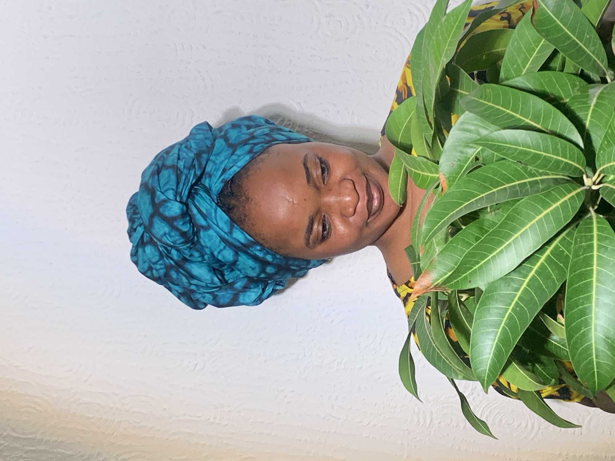 A person with brown skin and a dark-colored patterned headwrap is positioned against a white wall. The person's face is turned towards the camera, and they appear to be smiling gently. A vibrant green plant with elongated leaves partially obscures the right side of the frame. The background wall has a subtle textured pattern. The location is Barakin Akawo, Nigeria.