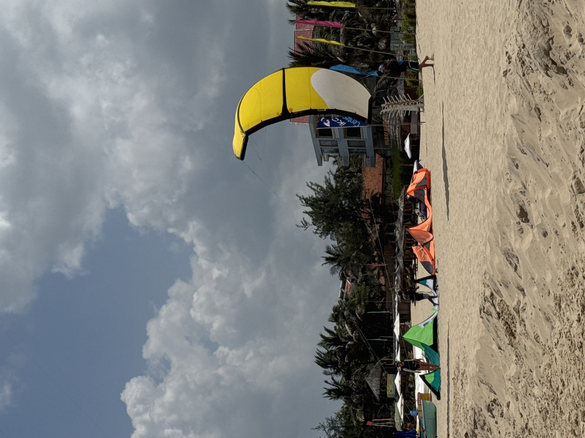 The image depicts a sandy beach in Mũi Né, Vietnam, under a partly cloudy sky. In the foreground, sand dunes are visible. The middle ground features several people and kitesurfing equipment. To the left, two individuals are preparing a green kite. Further to the right, an orange kite is partially laid out on the sand, with a person walking away from it. In the center-right, a large yellow and white kite is partially inflated and flying. In the background, palm trees line the shore, with buildings and colorful flags visible beyond them. A body of water is suggested by the presence of beach umbrellas and what appears to be a boat on the far left. The scene suggests an active kitesurfing or windsurfing spot.