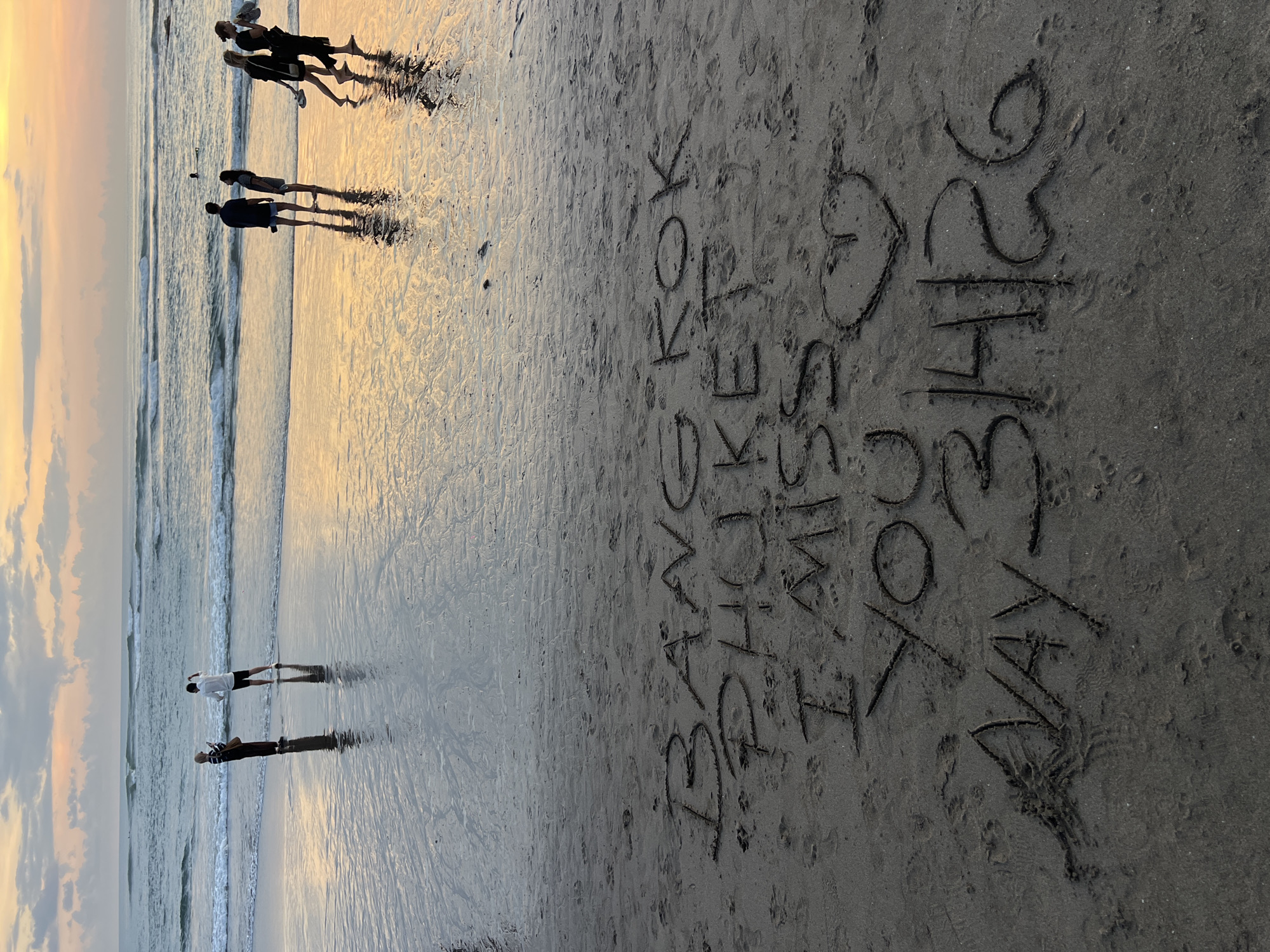 This photograph depicts a beach scene in Canggu, Indonesia. The photo appears to be taken at dusk, with warm light reflecting on the wet sand from the setting sun. In the foreground, text is written in the sand that states, "I <3 YOU MISS KOK BEACH MAY 31st 2026". Several people are present at the edge of the shore, with others a bit further into the water.