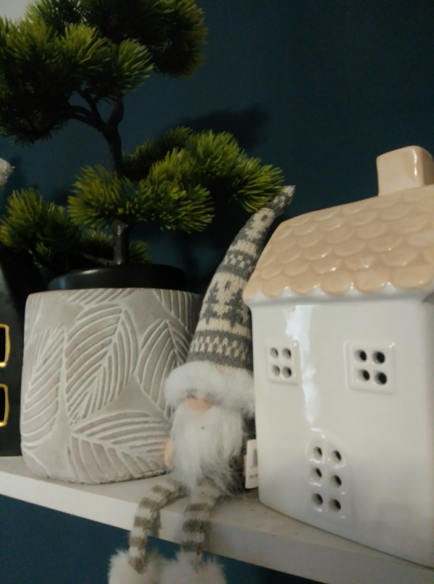 This image shows a shelf with decorative items. On the left is a potted artificial bonsai tree with green needles, set in a grey pot with a leaf pattern. To the right of the pot is a gnome figurine with a grey and white patterned hat and striped legs. Next to the gnome is a white ceramic house-shaped lantern with a tan, scaled roof. The house has square window openings. The items are arranged on a white shelf against a dark teal background. The scene is likely a home interior, possibly in Wedel, Germany, but the image itself does not provide specific location markers.