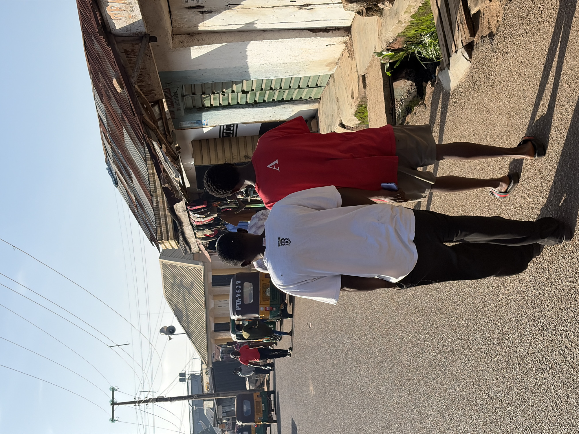 Two young men walk down a dusty street in Jos, Nigeria, with their backs to the camera. The man on the left wears a white t-shirt with a black logo on the back and black pants. The man on the right wears a red t-shirt with a white letter "A" on the back and khaki shorts. They walk on a paved road next to shops with corrugated metal roofs. The shops appear to be open for business, with goods displayed outside some of them. In the background, several motorized rickshaws, known as "keke napep" in Nigeria, are parked and some appear to be in motion. Overhead power lines crisscross the sky, and a utility pole with loudspeakers is visible. The buildings are simple structures with white walls and various types of doors and windows. The overall scene depicts a typical street view in a bustling commercial area of Jos.