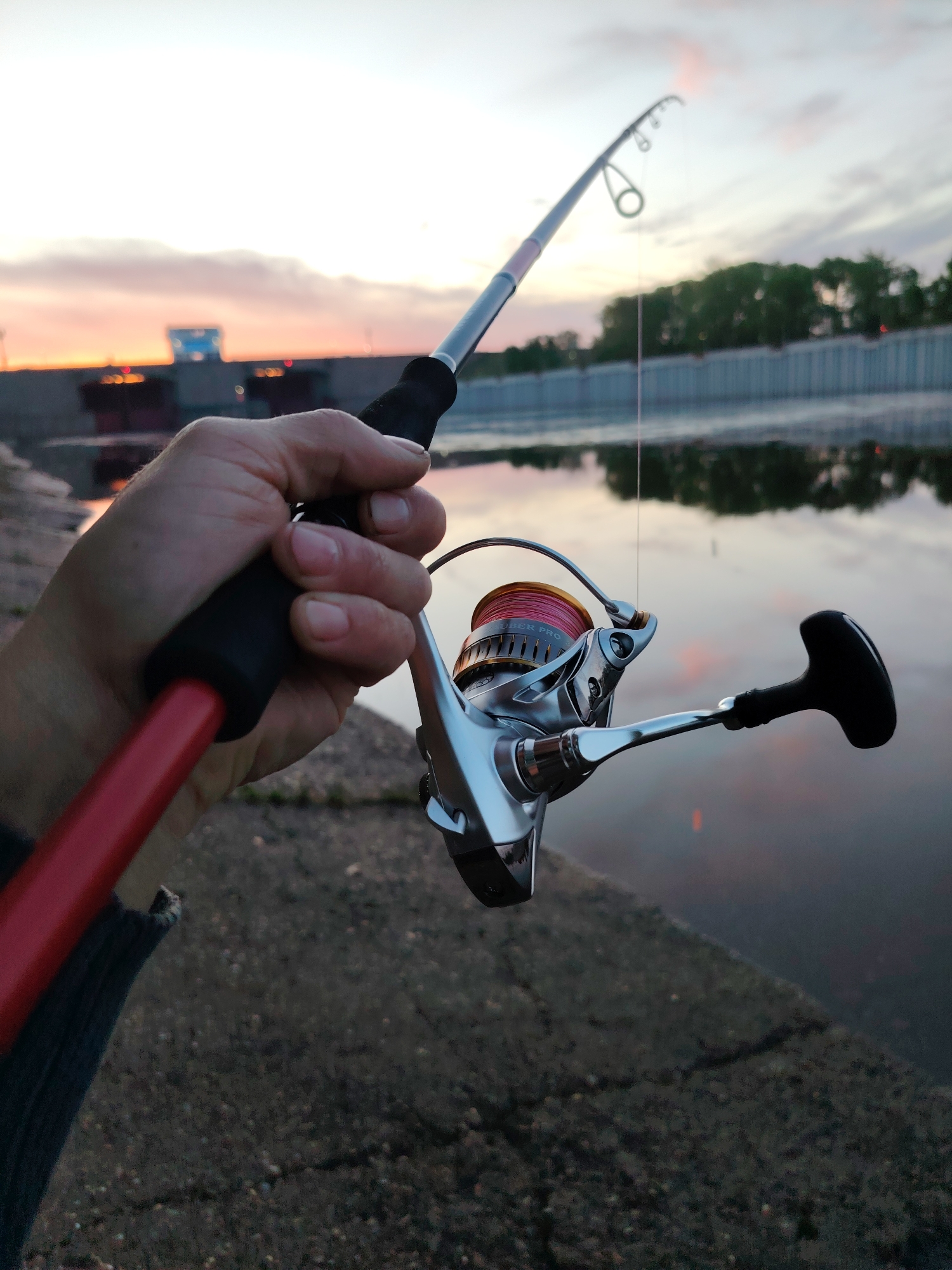 A close-up shot depicts a hand holding a fishing rod at what appears to be a waterfront location, likely the city of Chaikovsky, Russia, given the provided context. The fishing rod is red and black, with a silver reel wound with pink line. The line extends to the water's surface. The background features a canal or waterway with concrete walls. The water reflects the colors of the sky, which is a blend of pale oranges, pinks, and blues, suggesting either sunrise or sunset. Across the water is a structure that could be part of a dam or lock system, and some trees are visible in the distance. The foreground shows part of a concrete embankment or pier. A portion of a blue garment is present at the bottom left of the image.