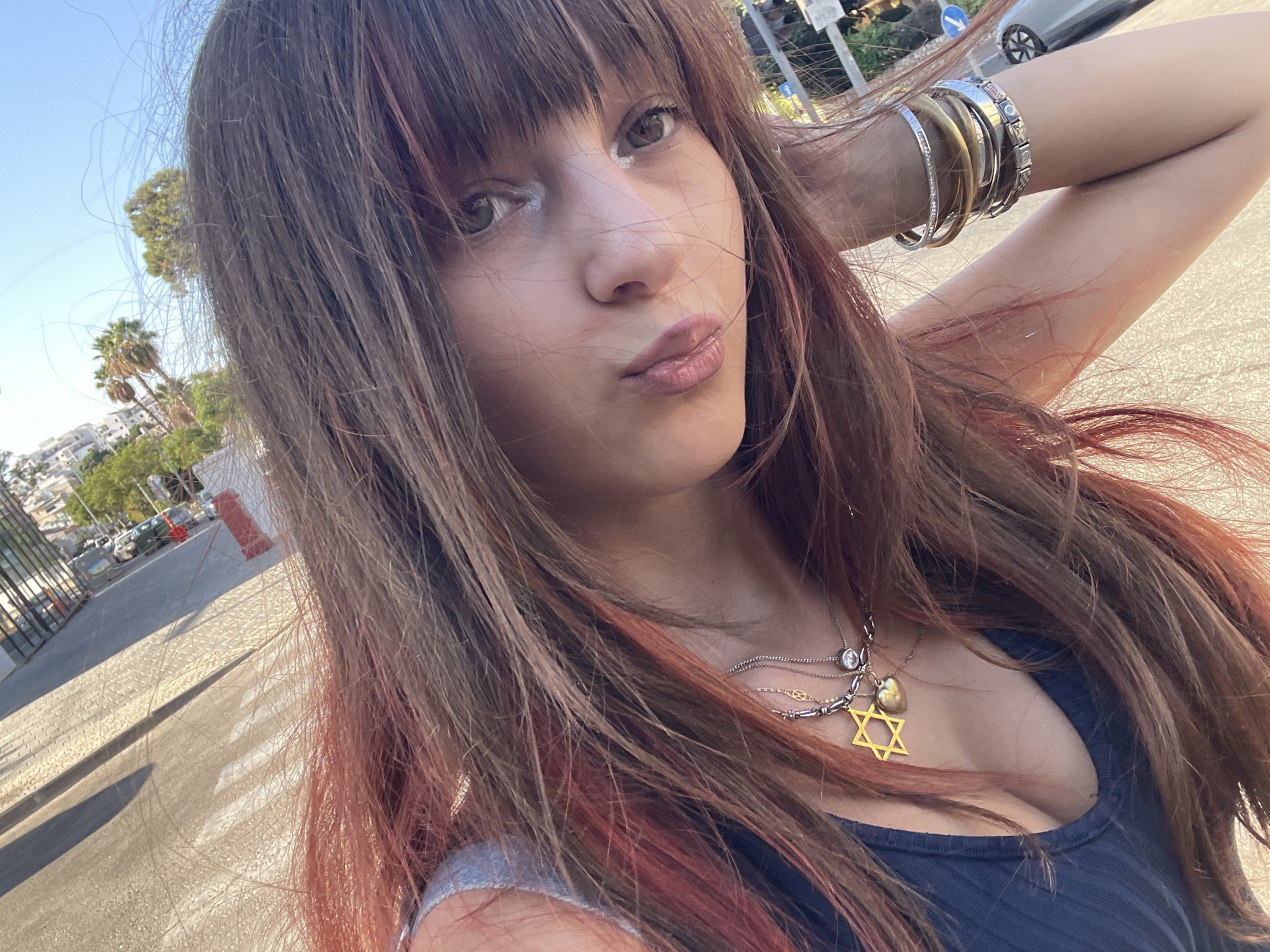 A young woman is captured in a selfie-style photograph, outdoors in a sunlit environment. She has long, reddish-brown hair with lighter streaks, styled with bangs. She is wearing a dark blue tank top and several necklaces, including a prominent yellow Star of David pendant and a heart charm. On her left wrist, she wears a stack of bracelets. Her makeup includes light eyeshadow and lip gloss. The background suggests an urban setting with a paved area, possibly a street or sidewalk, with some greenery and buildings visible in the distance. The lighting indicates it is daytime, likely late afternoon given the angle of the shadows. This scene is consistent with a public outdoor space in Petah Tikva, Israel.
